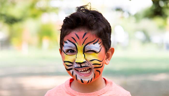 A smiling child with curly hair and a pink shirt has their face painted like a tiger, winking as they enjoy the outdoor fun at Canonsburg Live Music. The blurred green background adds to the vibrant atmosphere of this music festival.