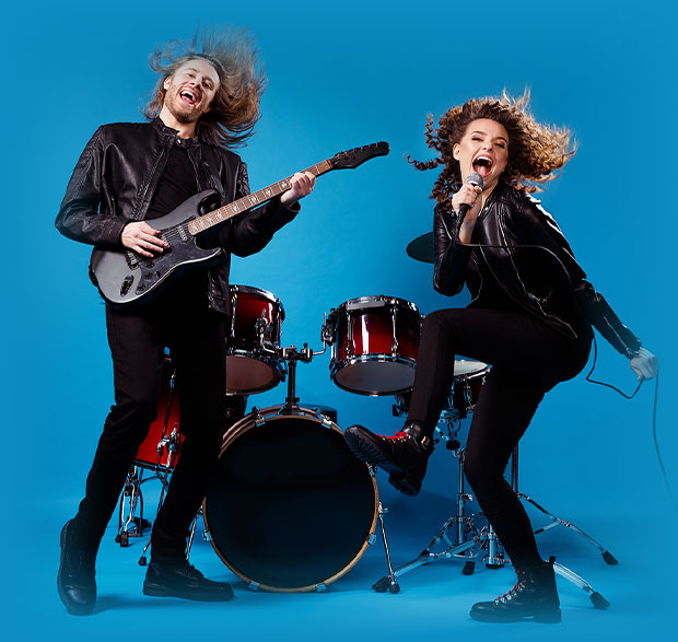 Two energetic musicians light up a blue background at Sunset Music Fest. One rocks an electric guitar while the other sings into a microphone. Both wear black outfits and leap in front of a drum set, capturing the spirit of live music.