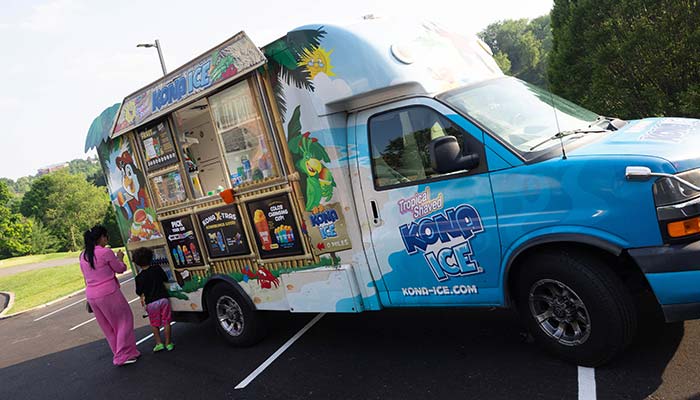 A blue Kona Ice truck with tropical-themed decorations is parked outdoors at the Sunset Music Fest. An adult and a child stand at the serving window, looking at the menu and ordering shaved ice treats while enjoying the festival atmosphere.