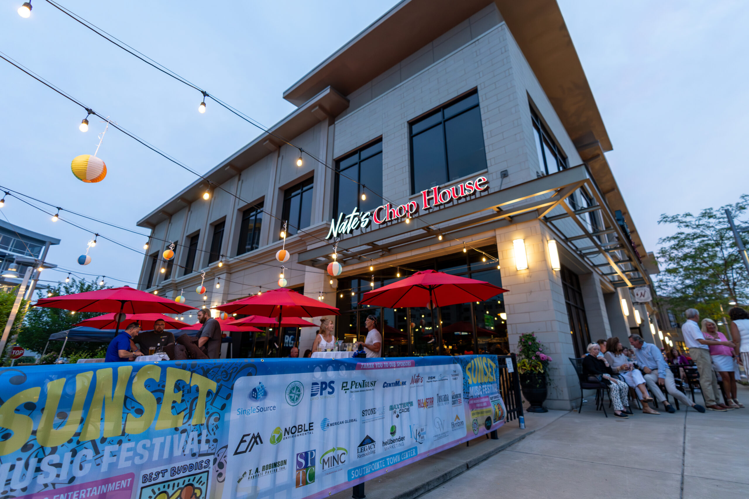 People dine outdoors under red umbrellas at Nate’s Chop House during sunset. String lights and colorful lanterns hang above, as a Sunset Music Fest banner in the foreground sets the scene for Canonsburg Live Music and local festival vibes.