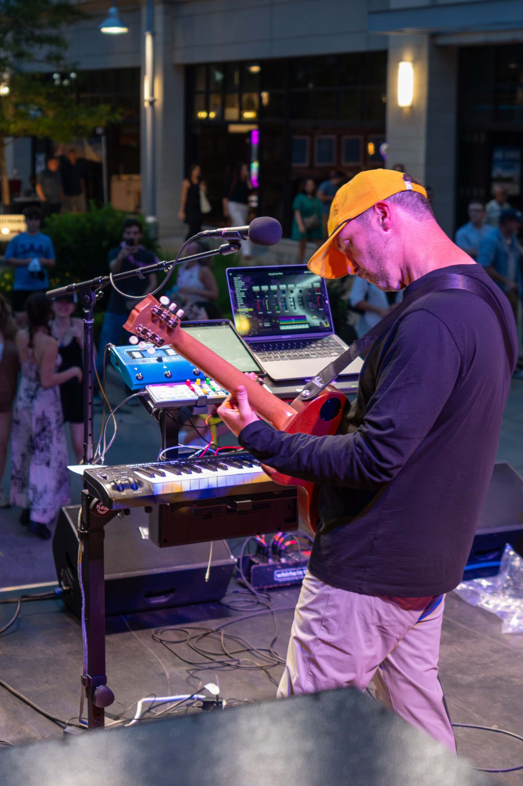 A man in a yellow cap and black shirt plays electric guitar on stage with music equipment and a laptop, while a crowd enjoys the outdoor performance at Canonsburg Live Music in the background.