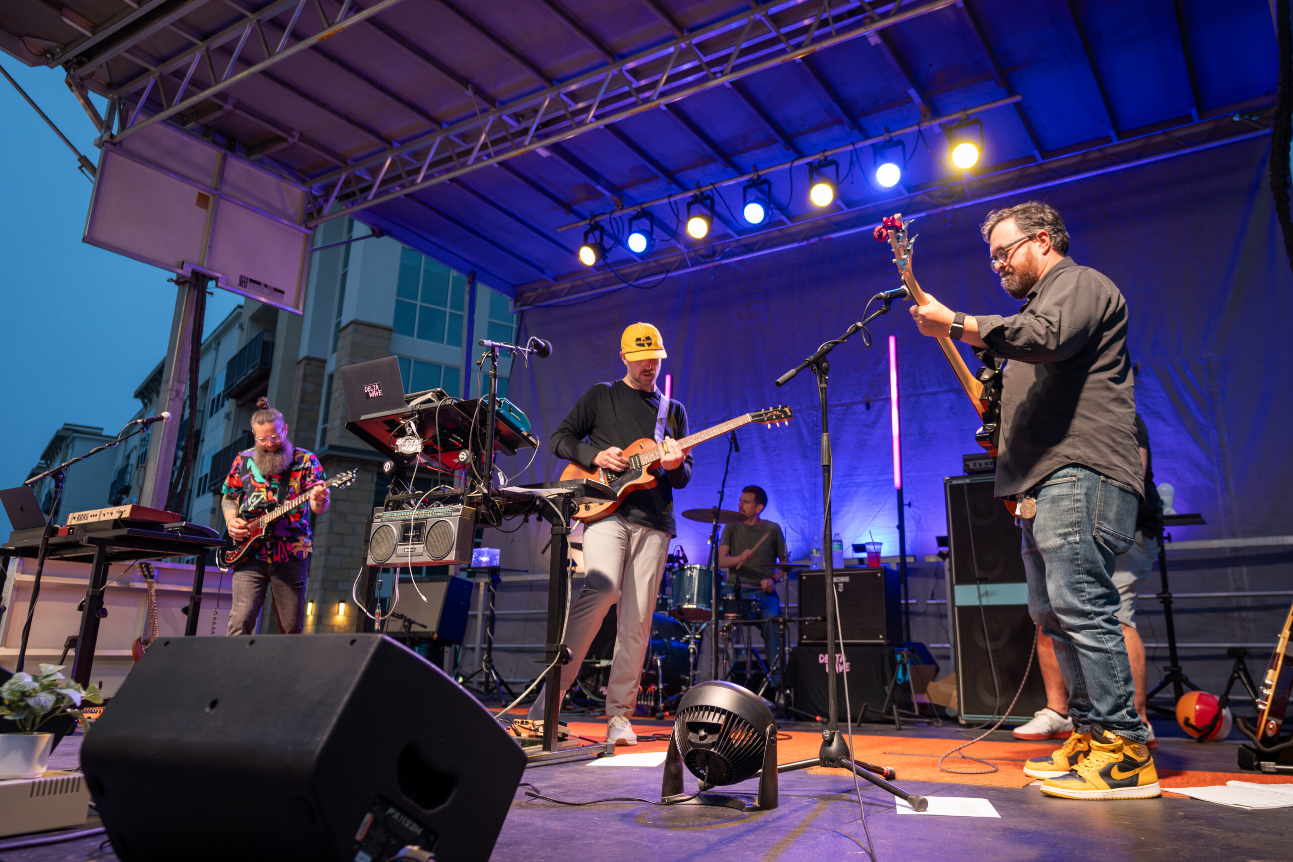 A band performs on an outdoor stage under colorful lights at the Canonsburg Live Music festival. Five musicians play electric instruments and drums, surrounded by equipment, with buildings visible in the background.