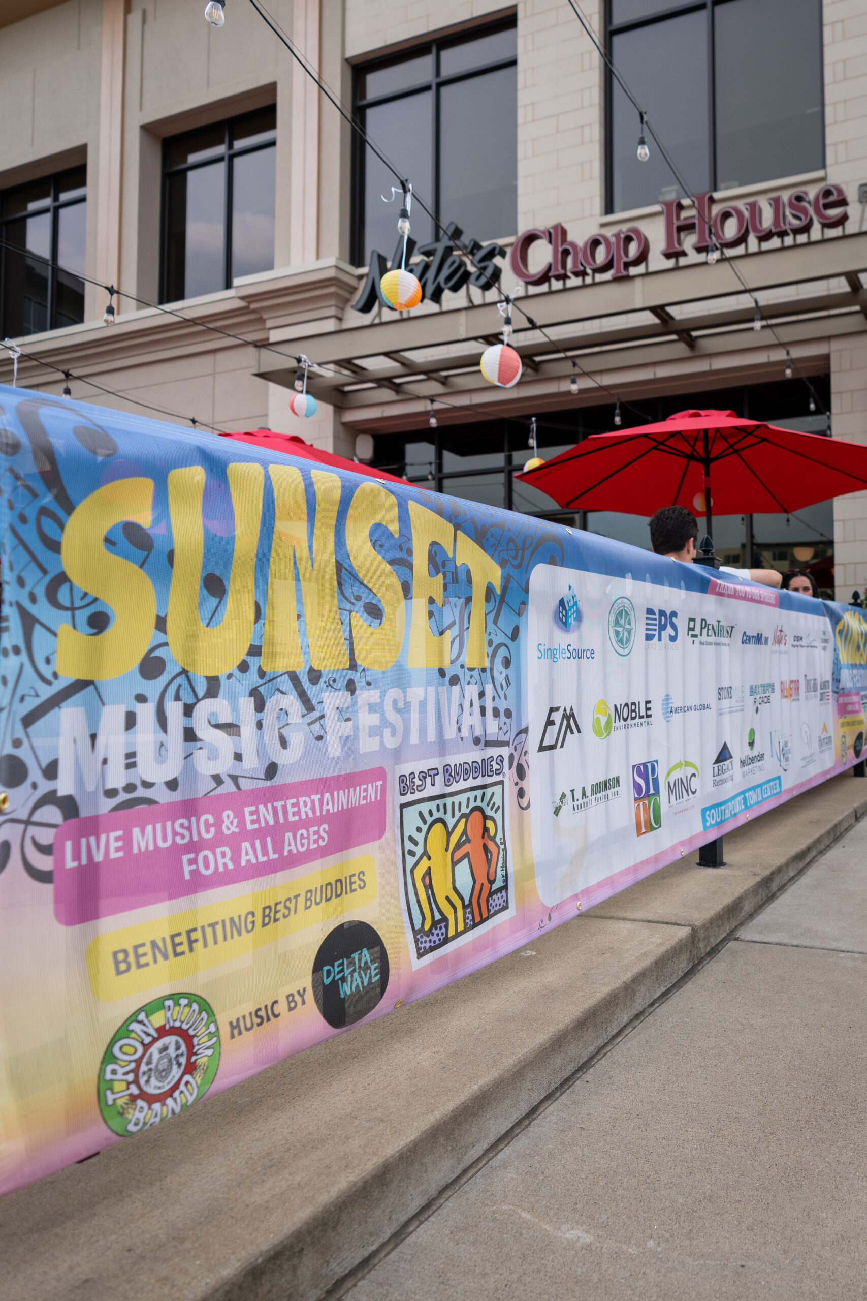 A colorful banner for the Sunset Music Fest hangs outside a building with a sign reading Chop House. Promoting Canonsburg Live Music and entertainment, the banner features sponsor logos, red umbrellas, and glowing string lights.