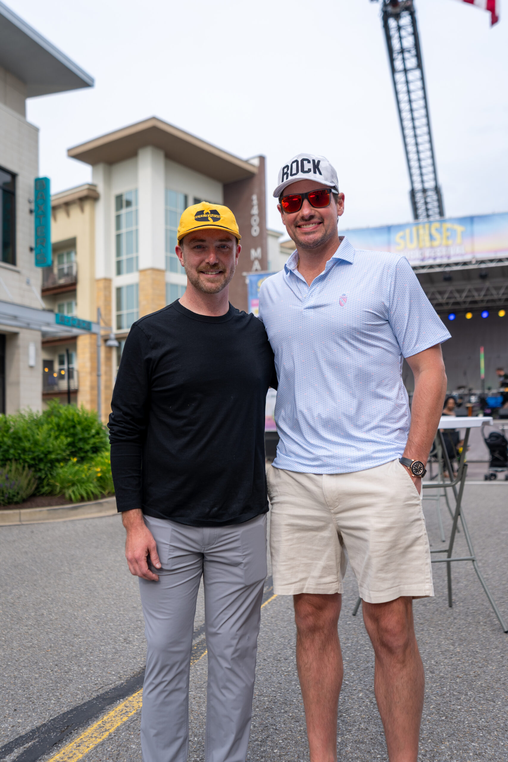 Two men stand side by side on a city street during the Canonsburg Live Music festival. One sports a yellow cap, black shirt, and gray pants; the other wears a white ROCK cap, sunglasses, blue polo, and khaki shorts. An outdoor stage appears in the background.