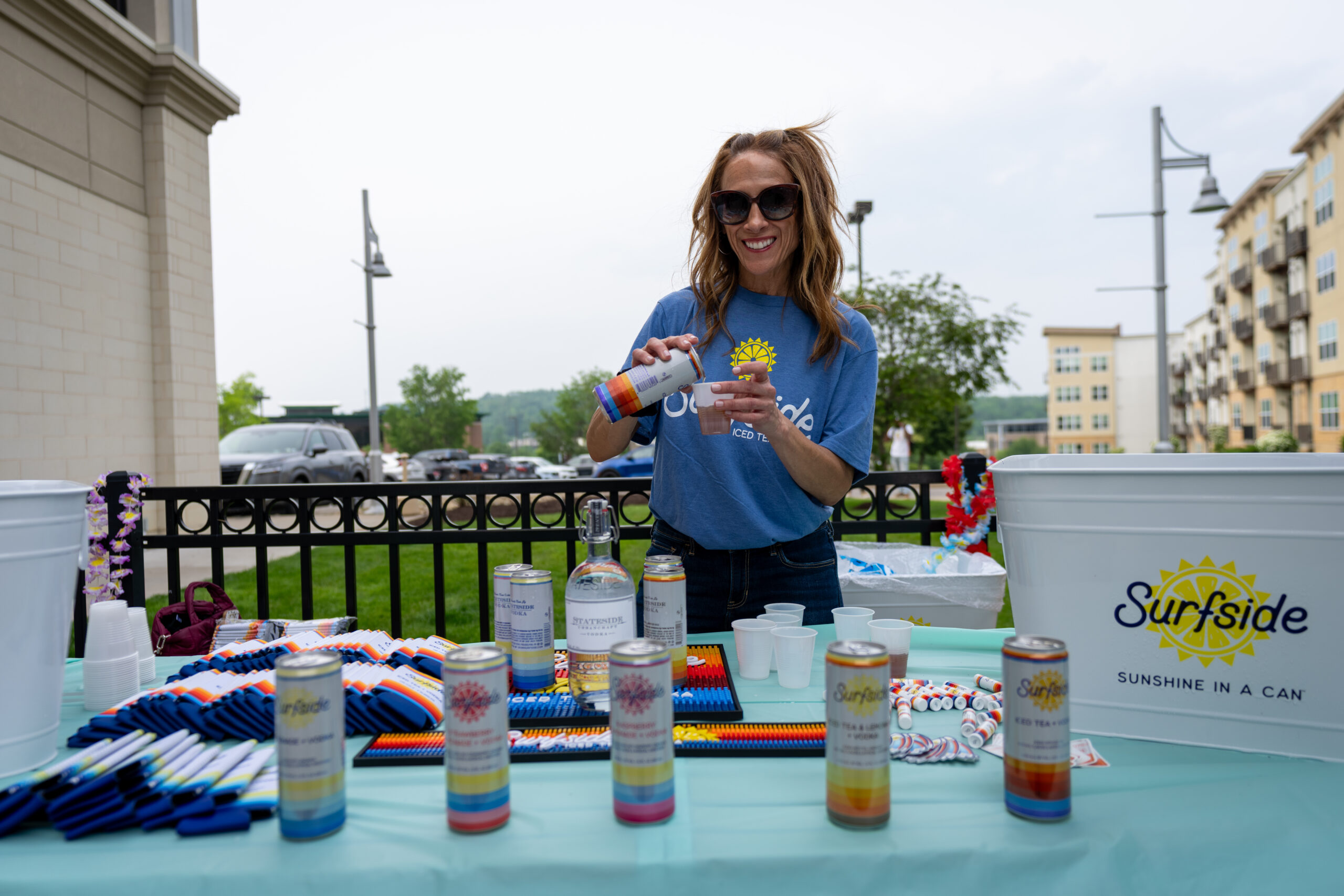 A smiling woman in sunglasses and a blue Surfside shirt pours a drink at an outdoor table with Surfside cans, cups, and a white cooler, enjoying the lively atmosphere of Canonsburg Live Music in front of a building and trees.
