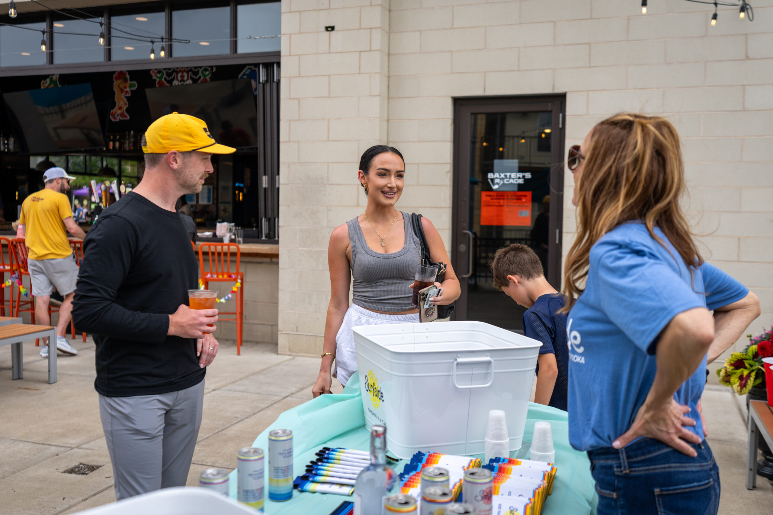 A group of people chat around a table with drinks and snacks at an outdoor event during Sunset Music Fest. One woman holds a drink while others stand nearby, and colorful cans and cups decorate the table. A restaurant with outdoor seating is in the background.