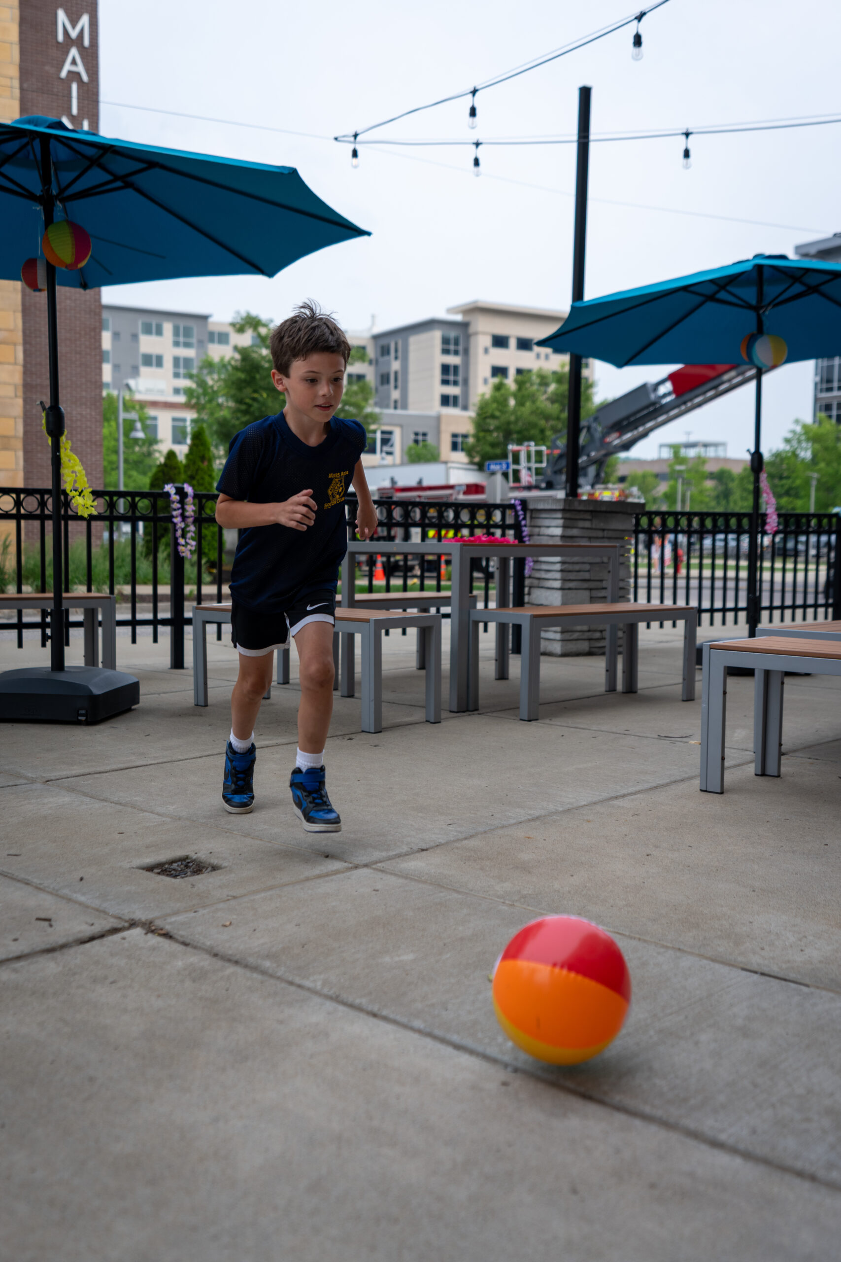 A young boy in a navy shirt and shorts runs outdoors towards a colorful beach ball on a concrete patio at the Sunset Music Fest, surrounded by tables, benches, and large blue umbrellas. Urban buildings and greenery are in the background.