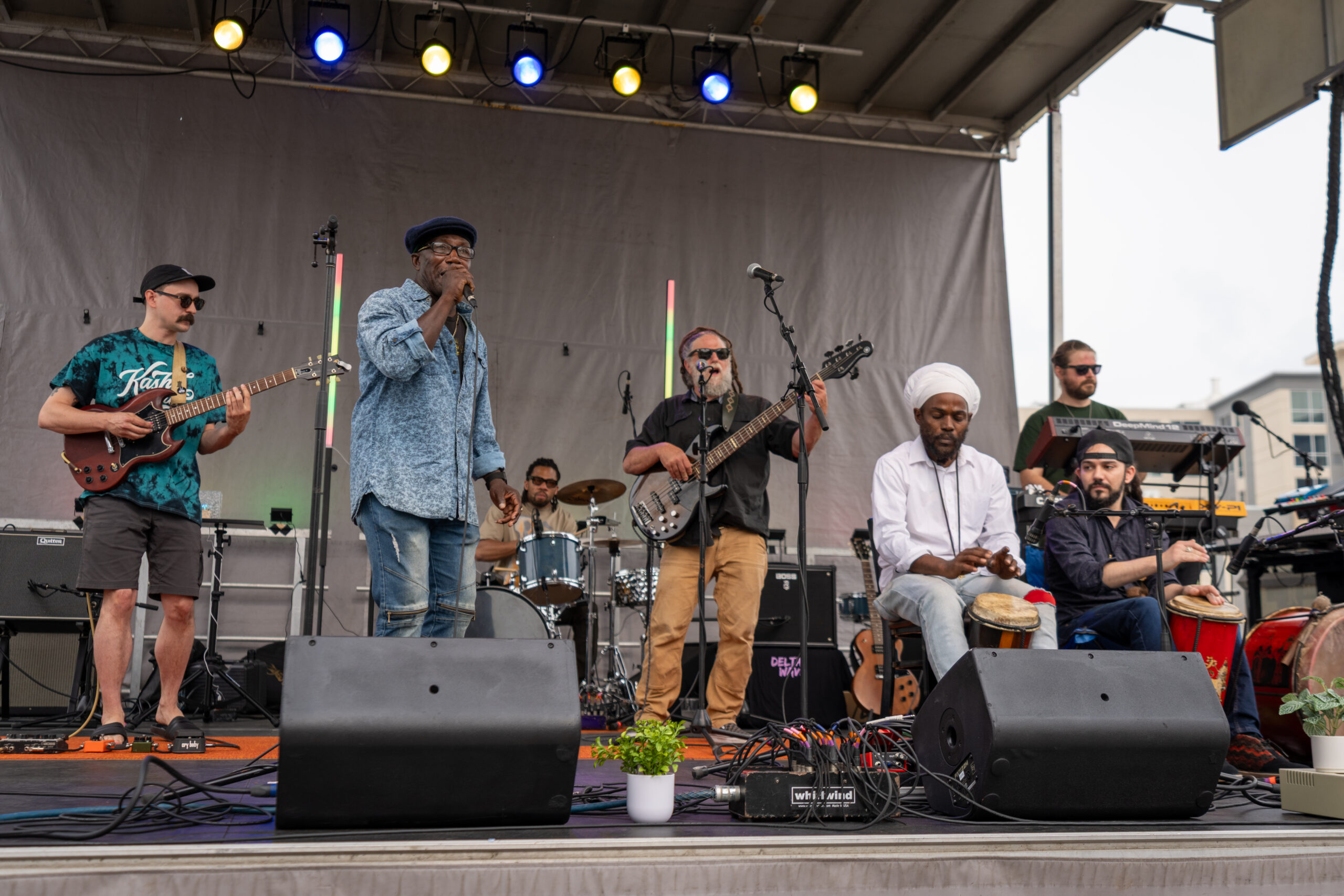 A diverse six-member band performs on an outdoor stage at Sunset Music Fest with guitars, drums, keyboard, and percussion. The lead singer stands at the center under colorful lights, while potted plants decorate the stage front.