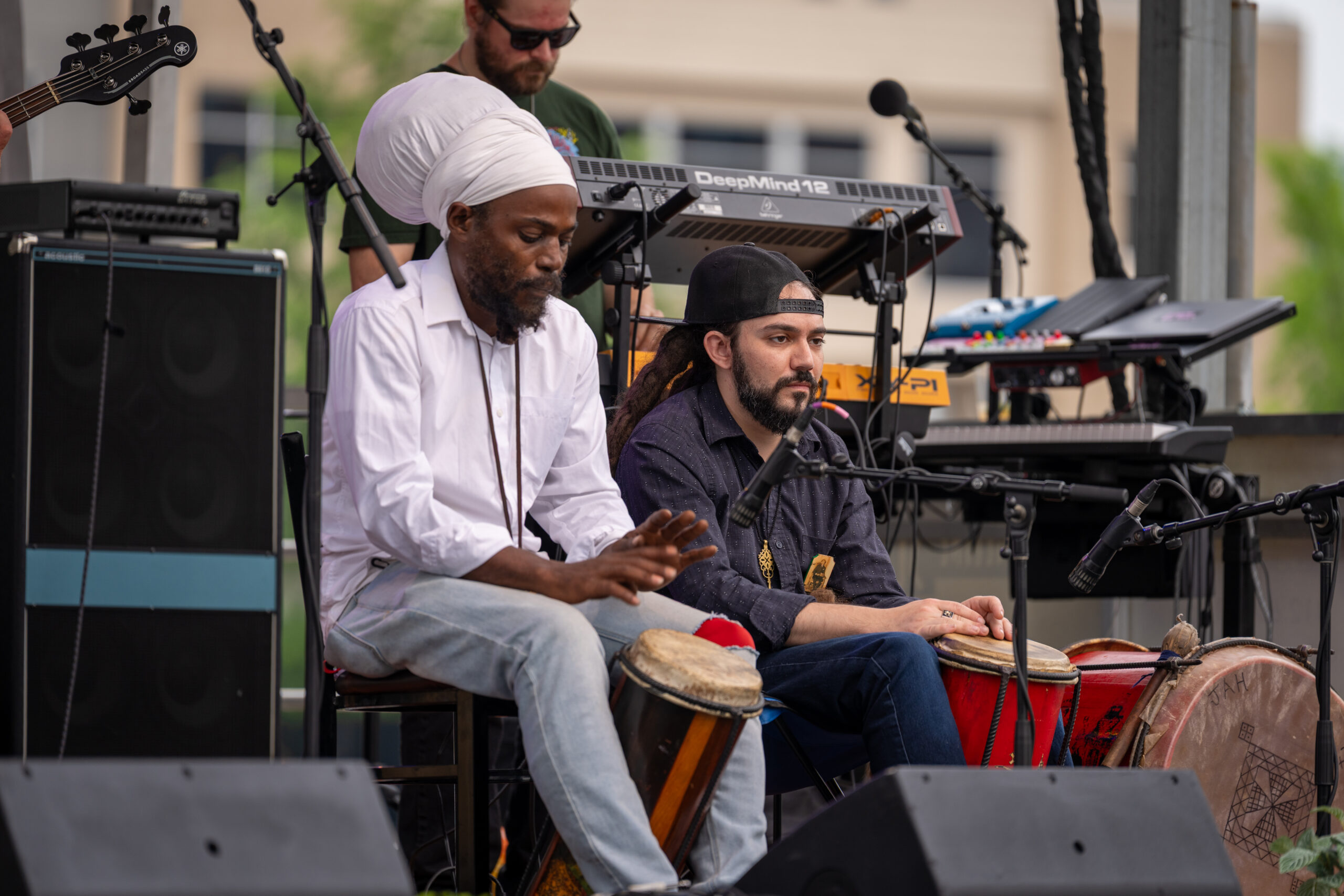 Two musicians sit on stage playing hand drums during an outdoor performance at Sunset Music Fest, with keyboards and other equipment visible behind them. One wears a white headwrap and shirt; the other has a black cap and long hair.