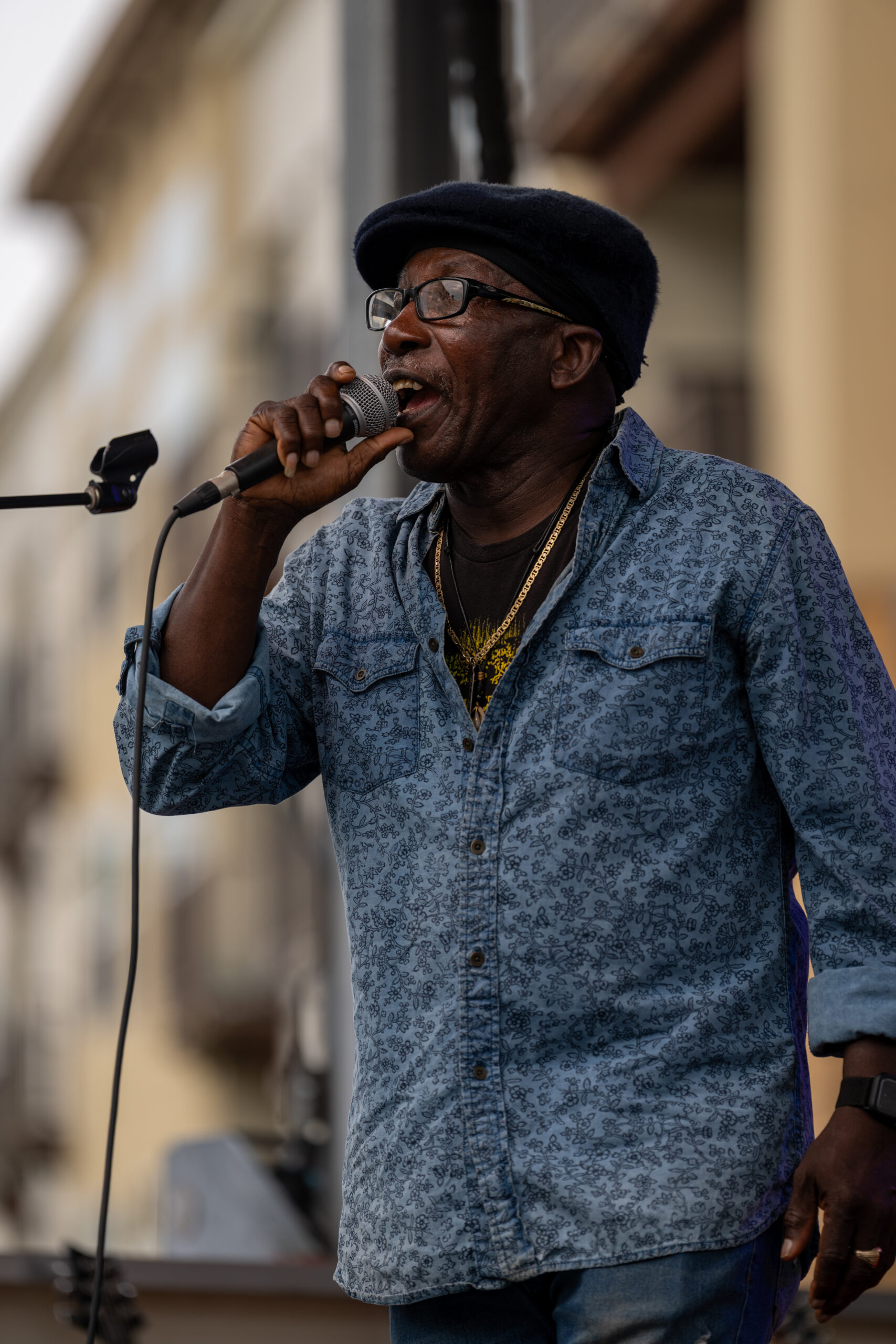 A man wearing a blue patterned shirt, glasses, and a dark cap passionately sings into a microphone on an outdoor stage at the Sunset Music Fest.