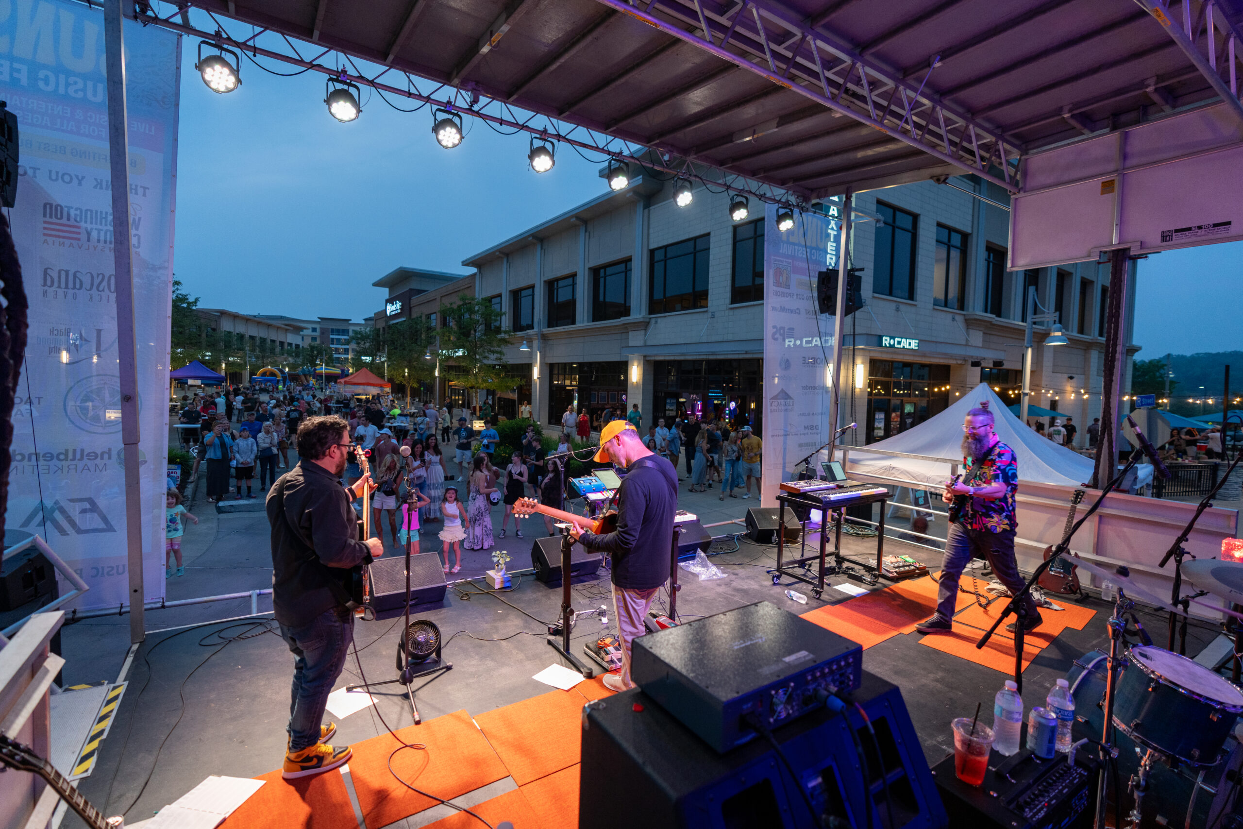 A band performs at Sunset Music Fest on an outdoor stage at sunset, with a lively crowd gathered in front of shops and restaurants under a blue sky. Stage lights shine on the musicians as people enjoy the live music.