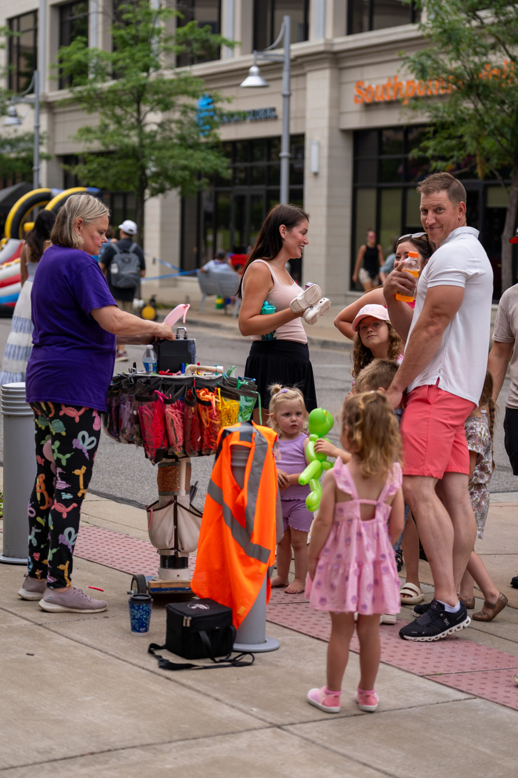 A group of adults and children gather around a street vendor selling colorful umbrellas on a city sidewalk during the Sunset Music Fest. A woman in purple stands by the stall, while kids and adults interact and smile. Storefronts and trees line the lively scene.