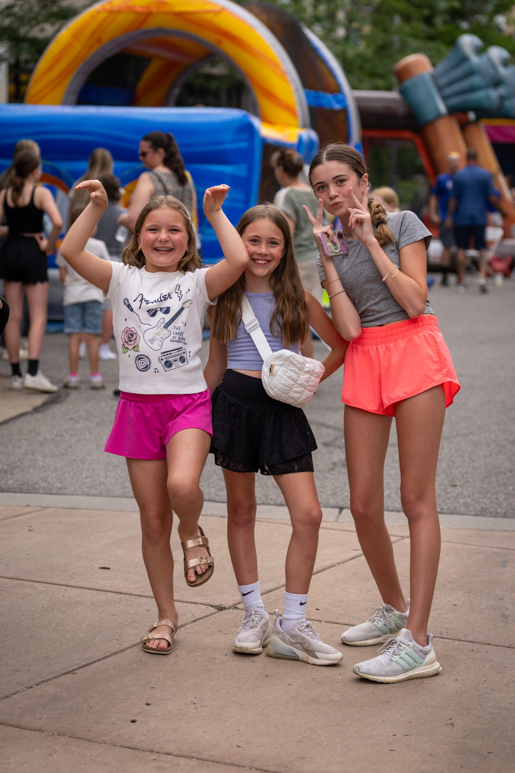 Three smiling girls pose playfully on a sidewalk at an outdoor event, with colorful inflatables and other people in the background. Dressed in casual summer clothes, they enjoy the lively atmosphere of the Sunset Music Fest.