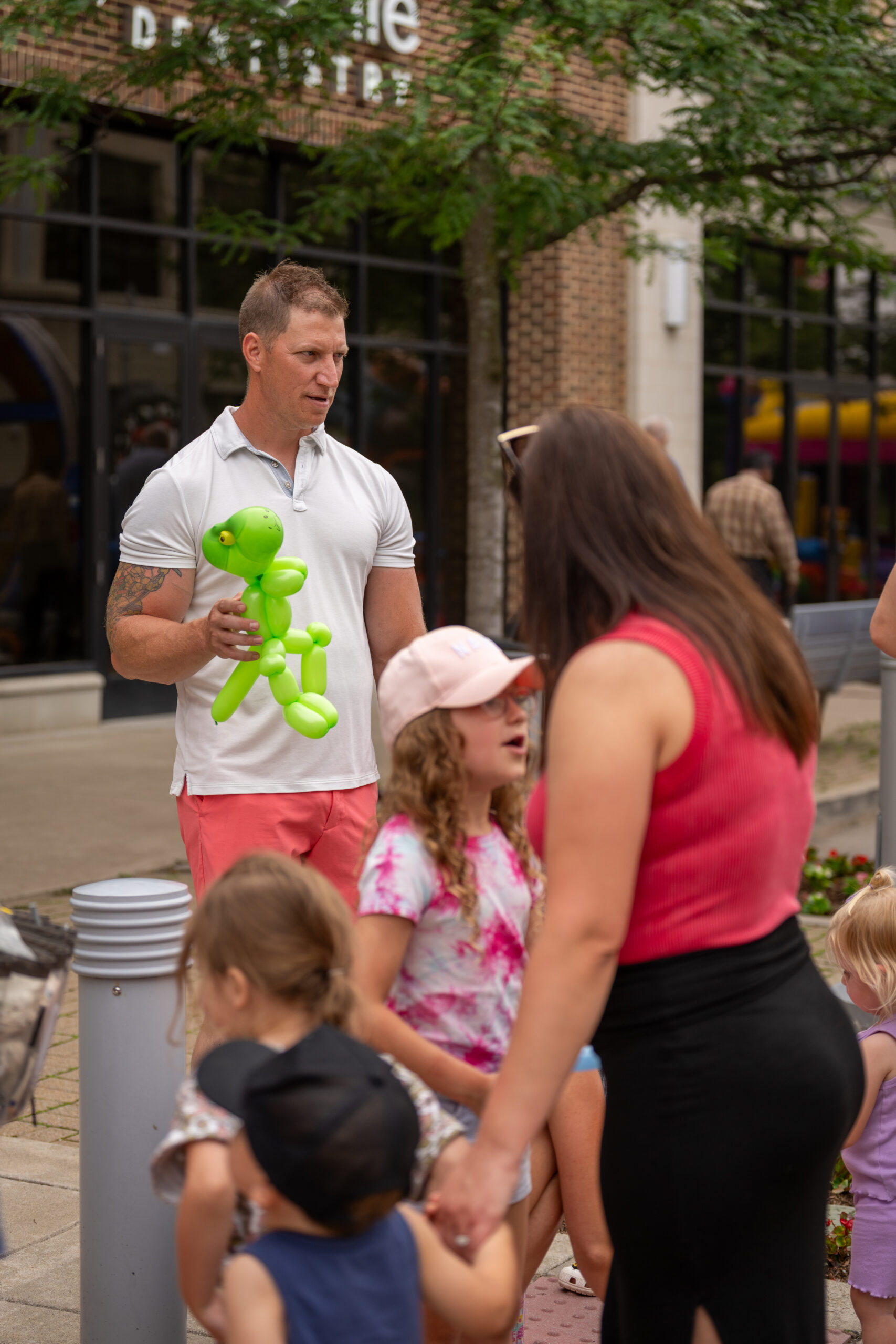 A man holding a green balloon animal stands outside near a group of children and adults, including a woman in a red top, on a lively street lined with trees and shops during the Sunset Music Fest.