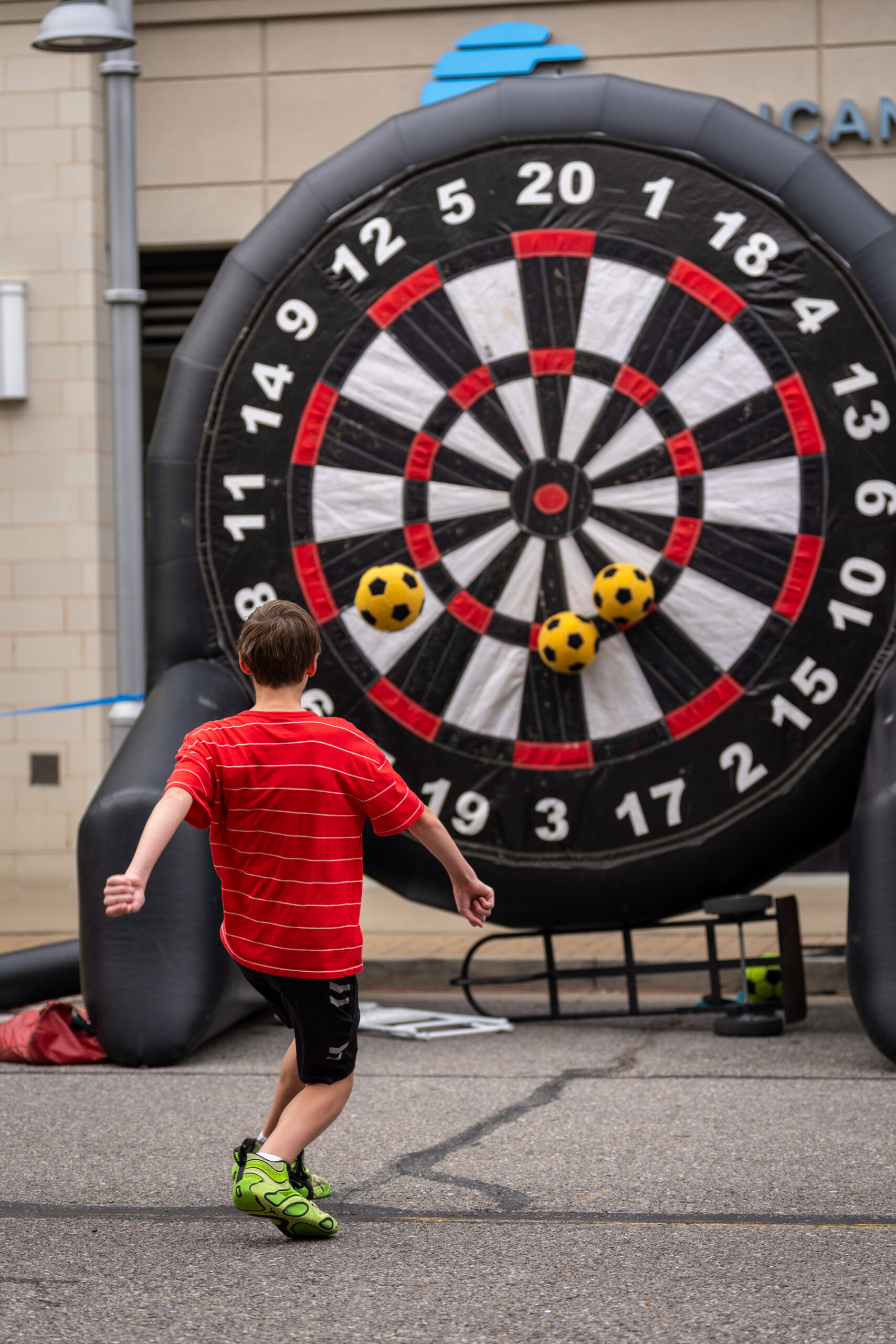 A boy in a red striped shirt and green shoes kicks a soccer ball toward a giant inflatable dartboard outdoors at Sunset Music Fest, with two soccer balls already stuck to the board.