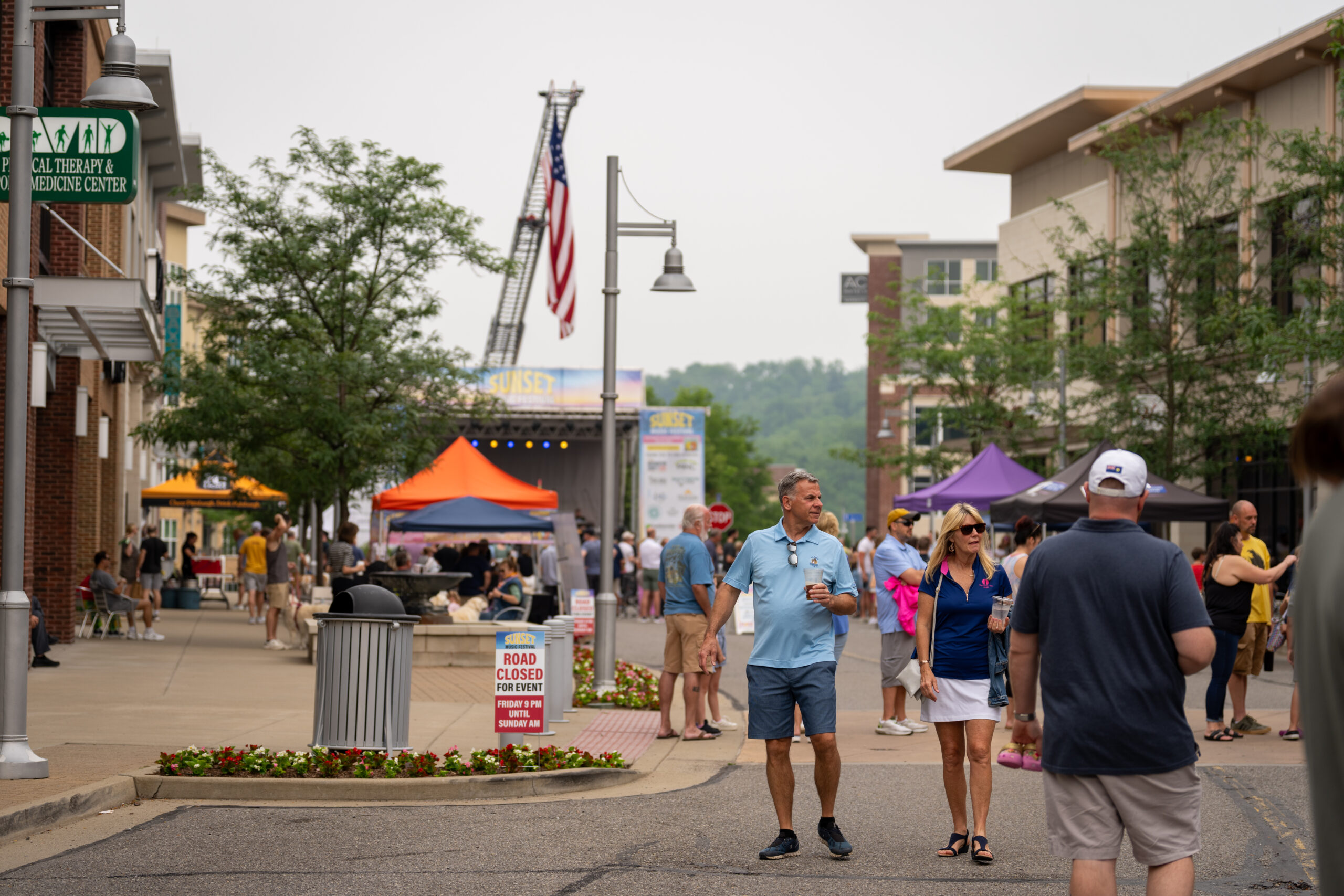 People stroll down a closed street at Sunset Music Fest, surrounded by colorful tents, a stage in the background, and an American flag. Trees and modern buildings line the lively scene at this Canonsburg live music event.