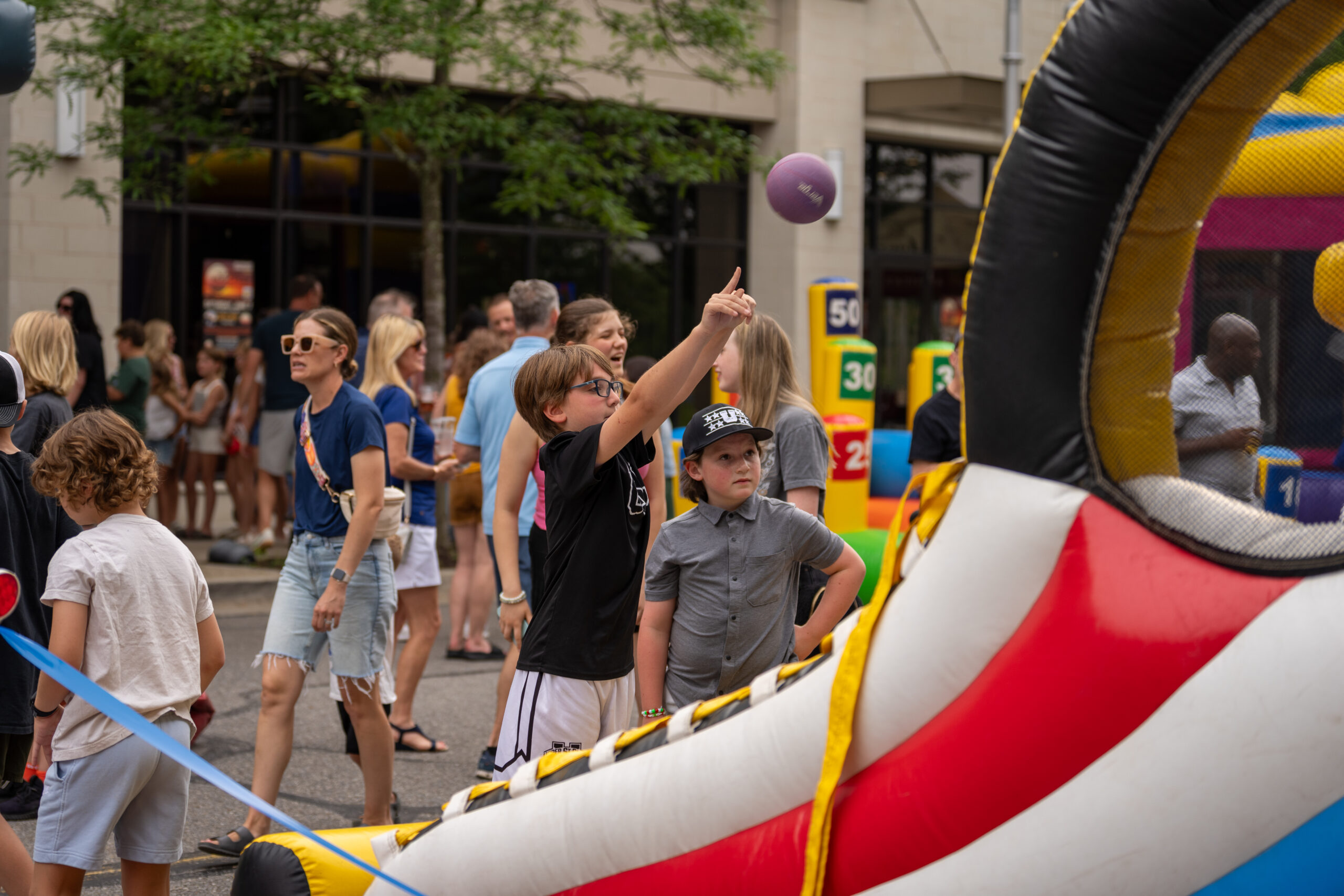 A boy throws a purple ball at a carnival basketball game while others watch and stroll around the outdoor street fair during the Sunset Music Fest. Colorful game booths and crowds create a lively backdrop in this festive atmosphere.