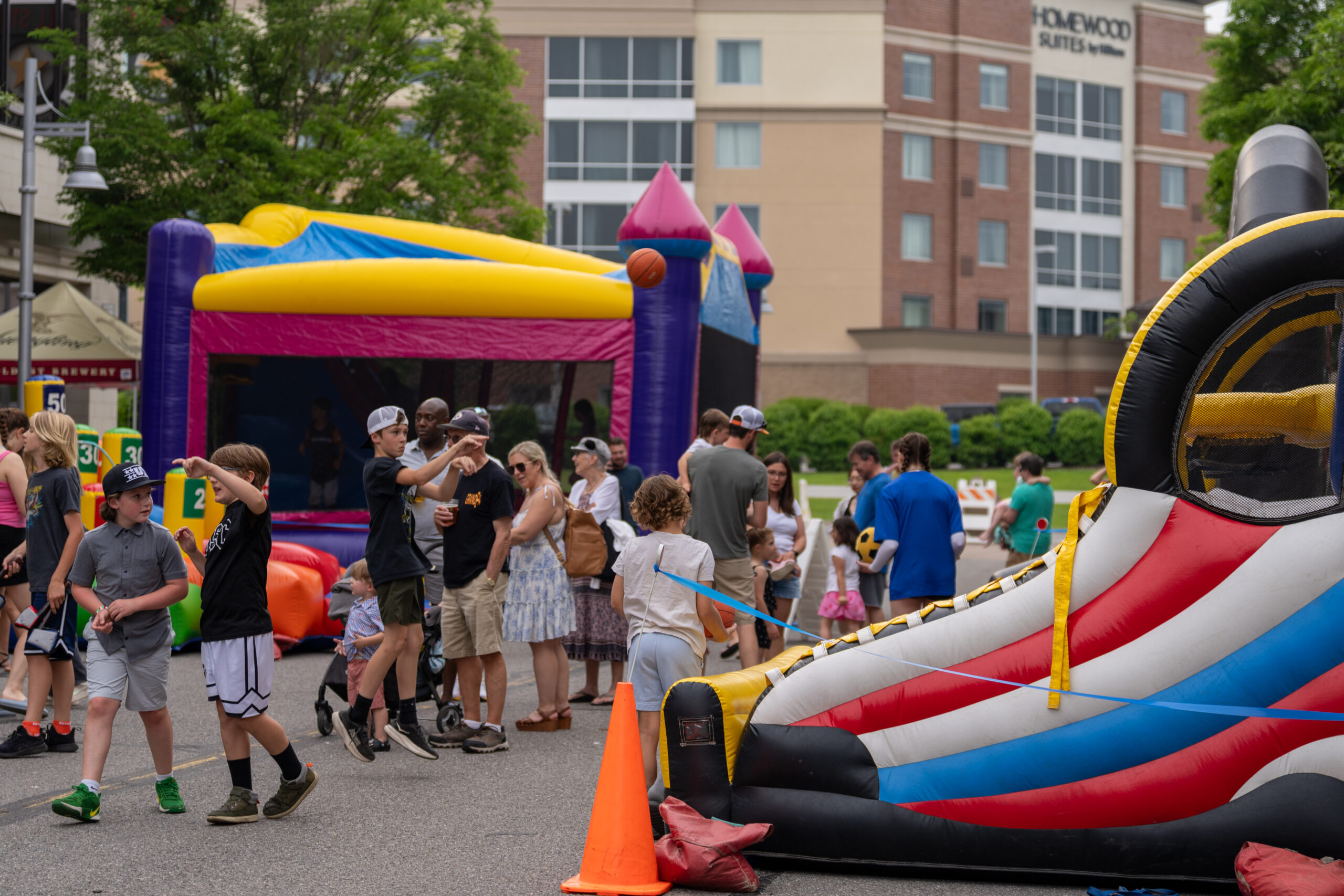 People gather at an outdoor event with inflatable bounce houses, games, and activities. Adults and children enjoy the Sunset Music Fest festivities on a closed street, with buildings and greenery in the background.