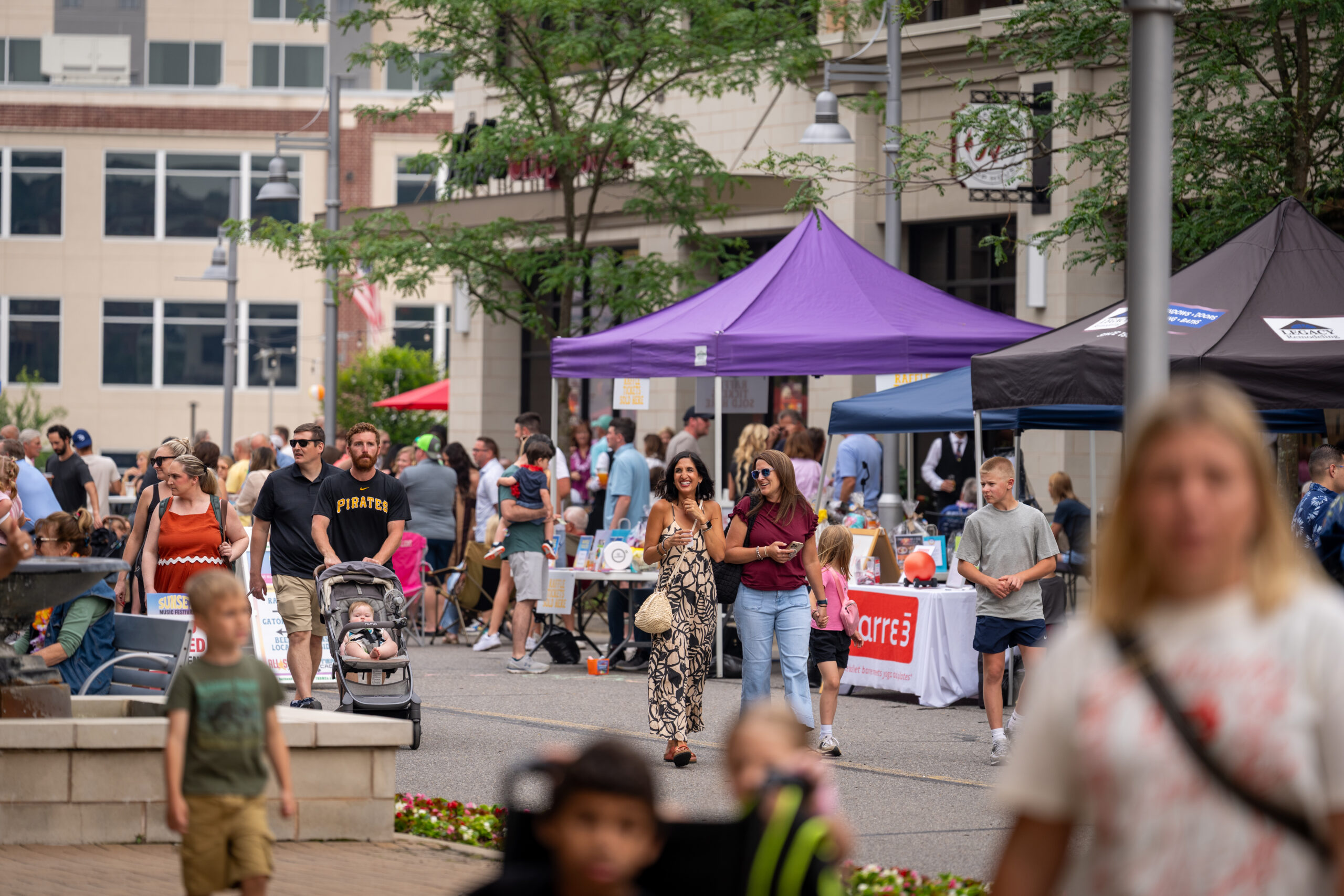 People walk through an outdoor market with vendor tents and tables, including a purple tent in the center, during Sunset Music Fest. Some people push strollers, others talk and shop. Trees and buildings line the background on a cloudy day.