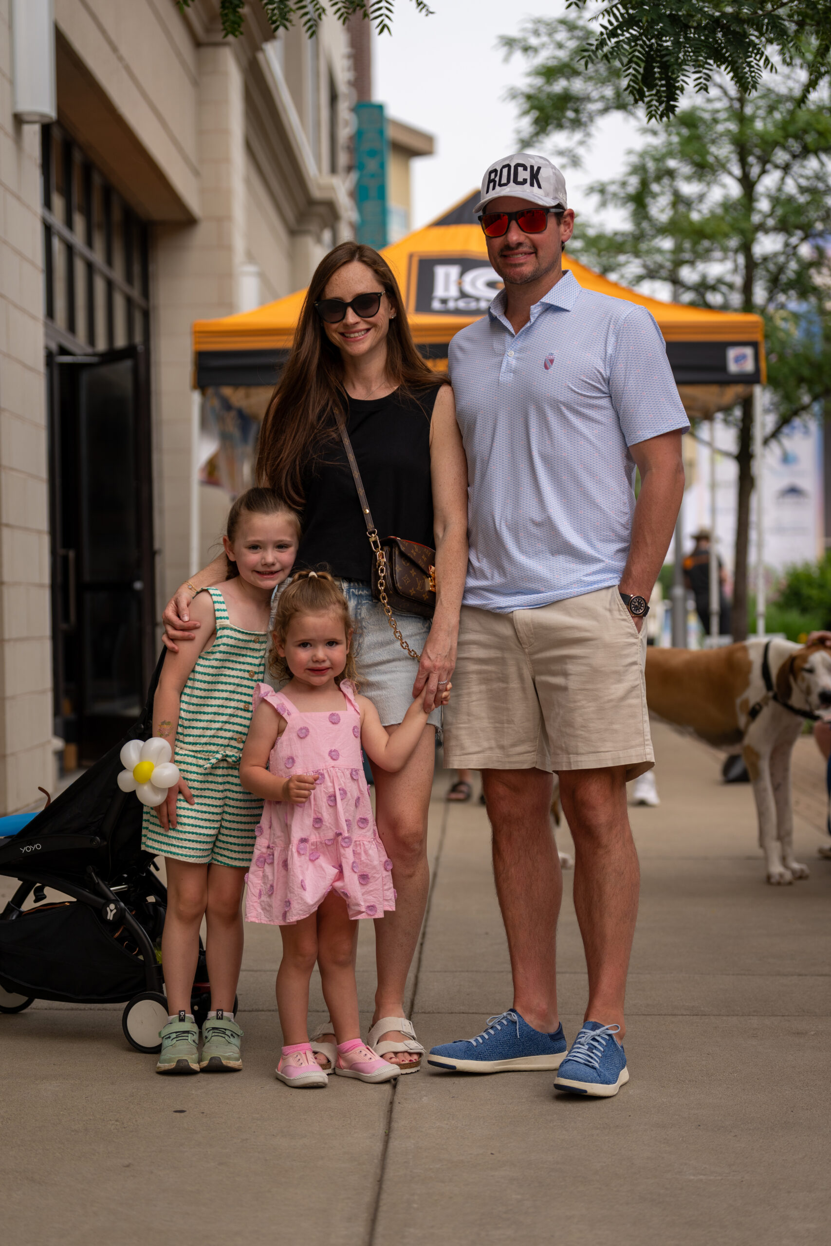 A family of four, two adults and two young girls in summer dresses, stands smiling on a sidewalk at Sunset Music Fest. The adults wear sunglasses, and a dog is in the background near a yellow tent at this lively music festival.
