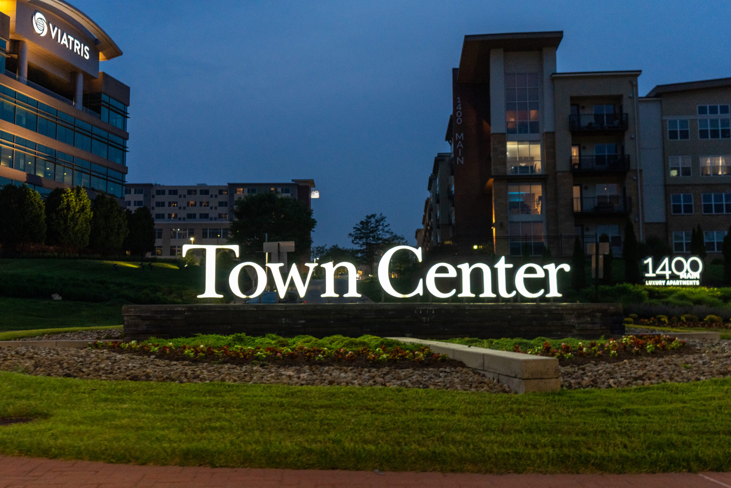 Illuminated Town Center sign at night, surrounded by grass and landscaping, with apartment buildings and 1400 Main and Viatris signs visible in the background—perfect for a Canonsburg Live Music night or the upcoming Sunset Music Fest.
