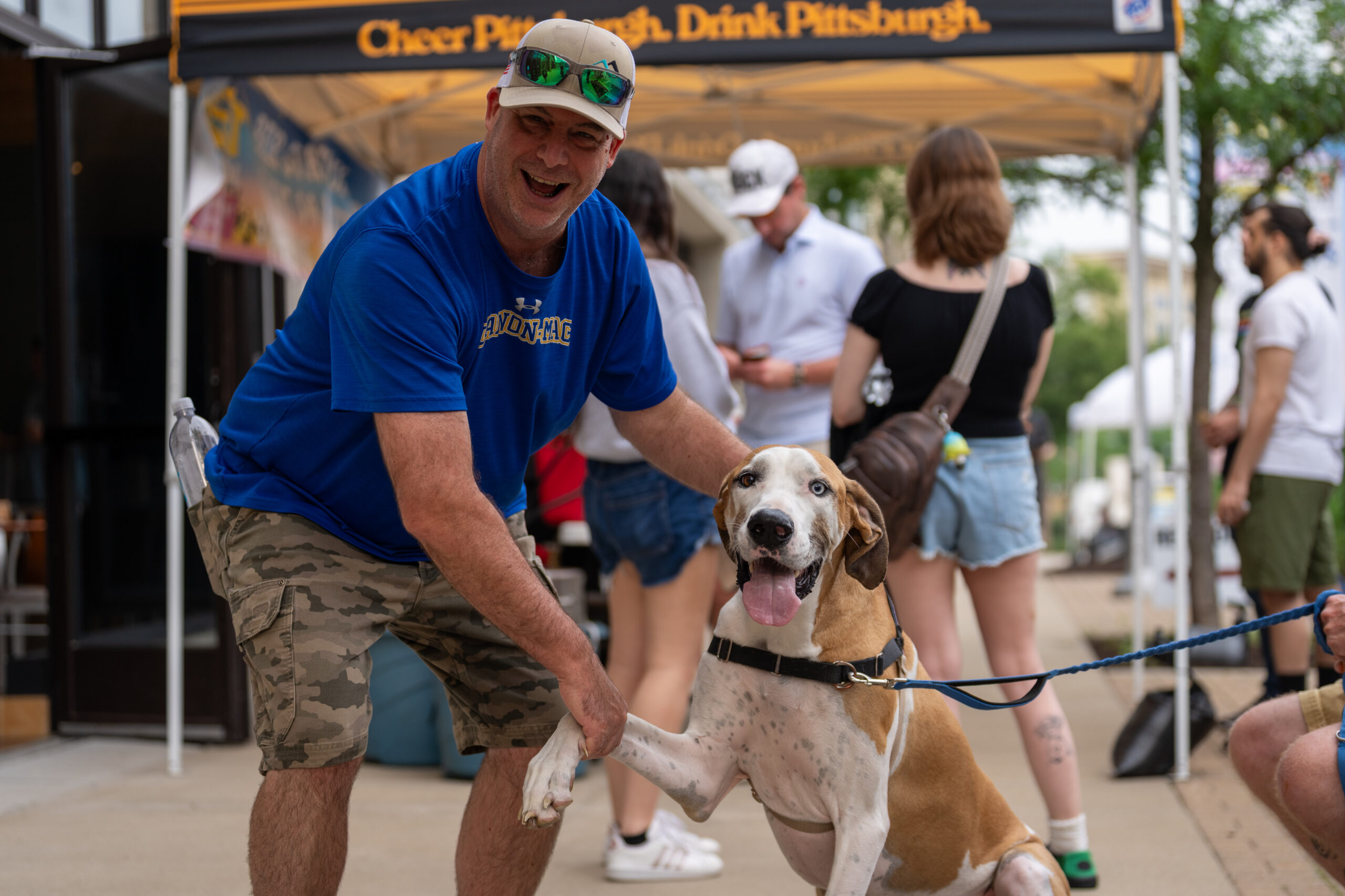 A man in a blue T-shirt and camo shorts smiles while holding the paw of a happy brown and white dog at an outdoor Sunset Music Fest, as people mingle in the background near a tent.