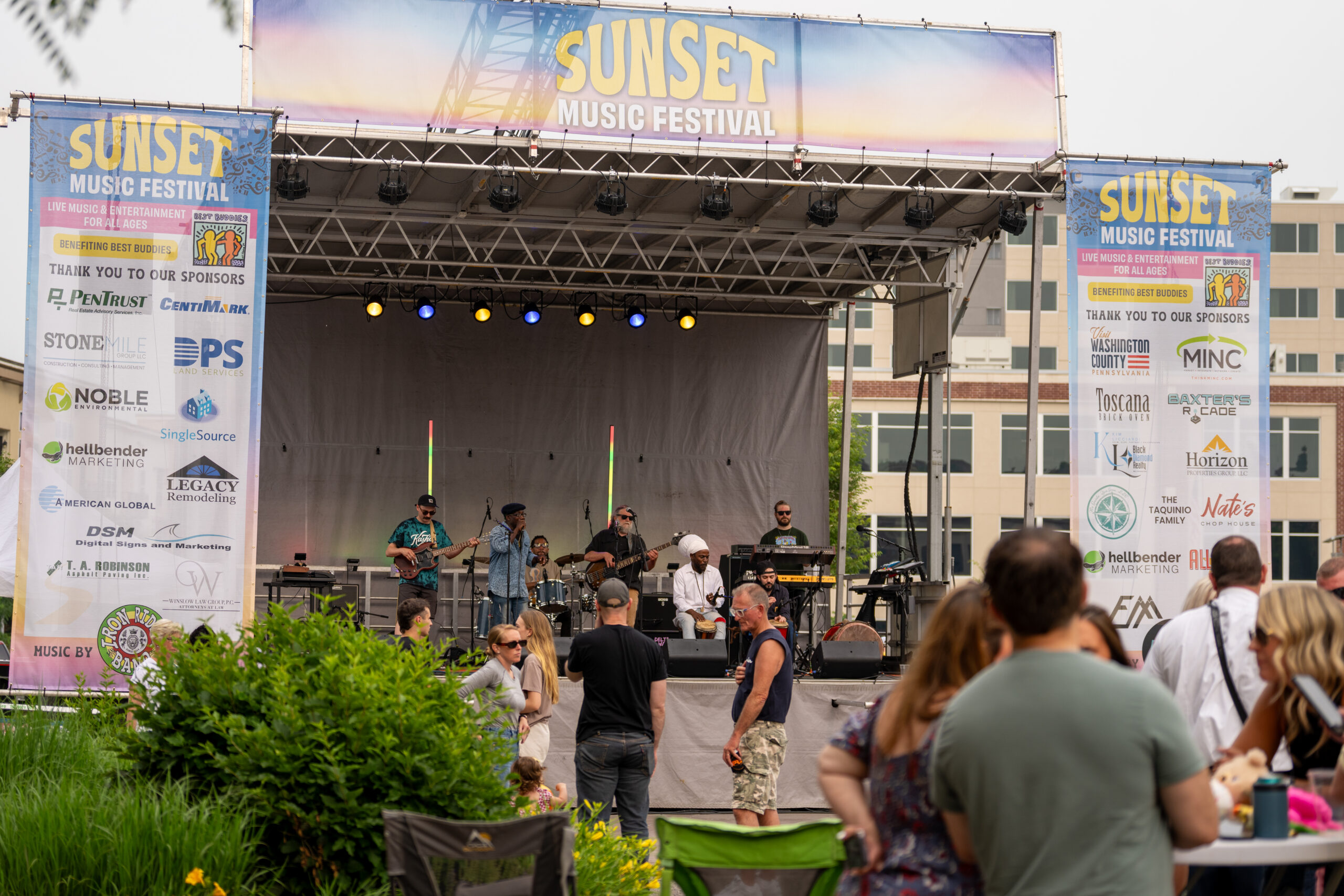 A band performs on an outdoor stage at the Sunset Music Fest, with sponsor banners on both sides. Audience members enjoy Canonsburg live music and socialize in front of the stage on a cloudy day.