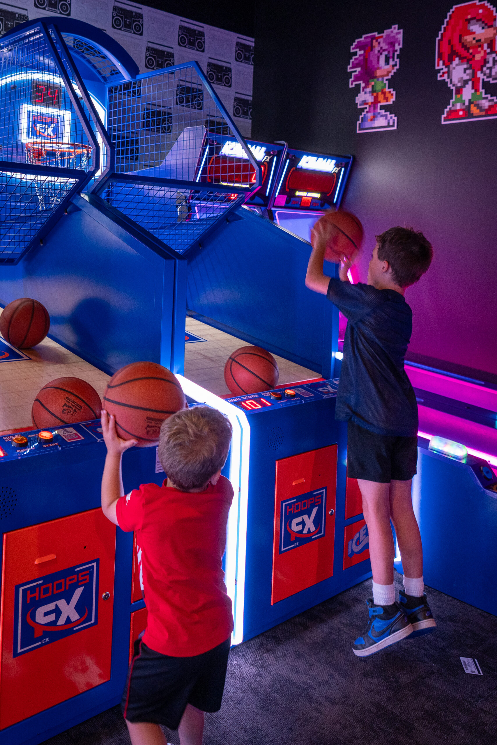 Two young boys play an arcade basketball game, aiming to shoot orange balls into hoops. The colorful setting, with pixel art characters and neon lights, captures the excitement of a Music Festival like Canonsburg Live Music.