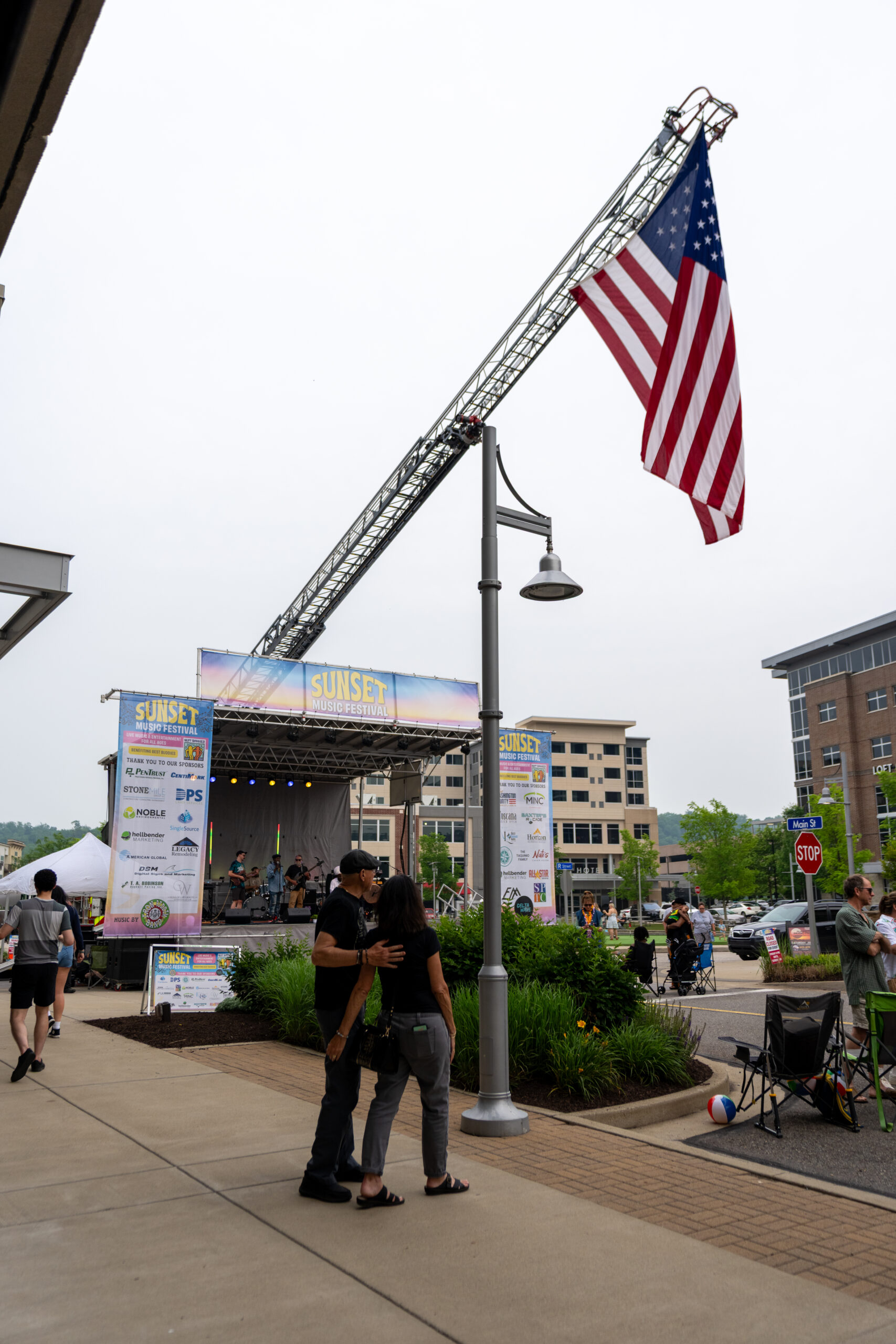 A couple stands close together near a festival stage under a large American flag hanging from a fire truck’s ladder at Canonsburg Live Music. Other people and buildings are visible in the background on this cloudy day.