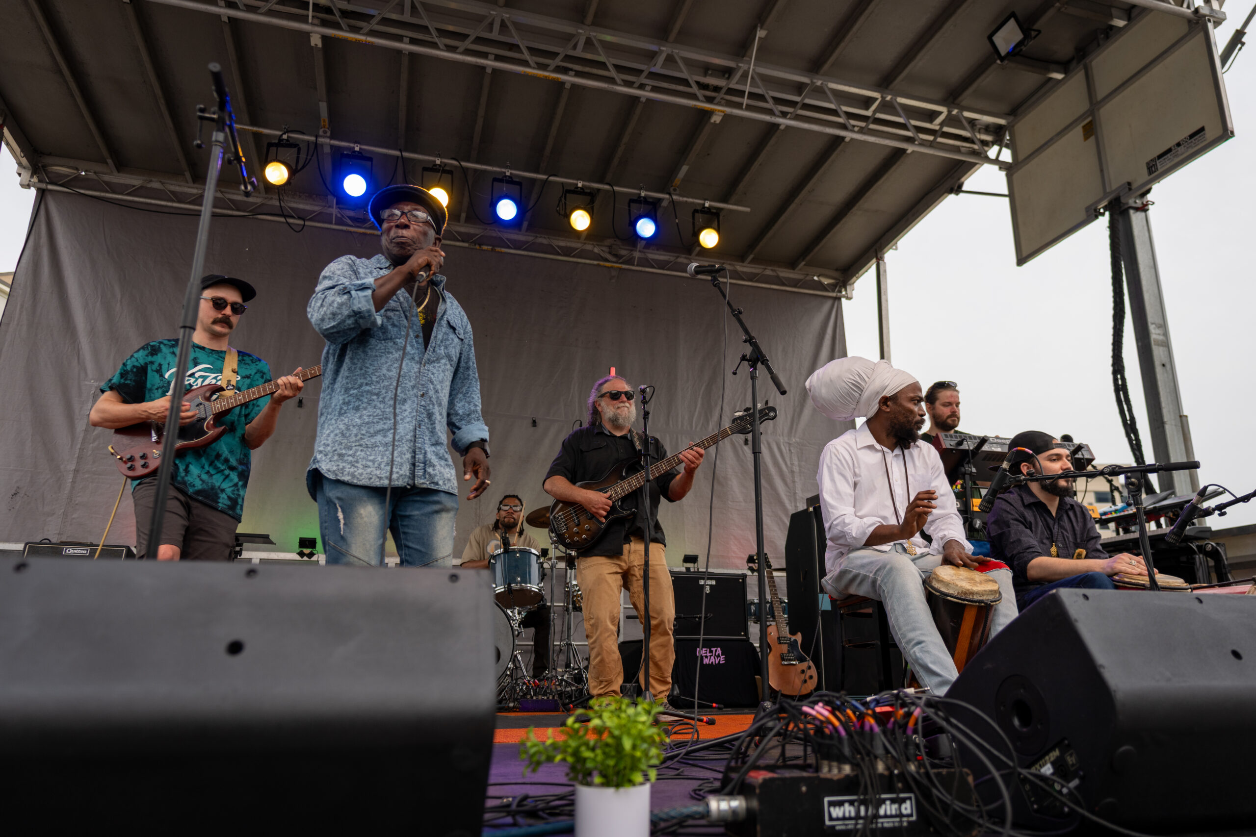 A band performs on an outdoor stage at Sunset Music Fest, featuring a singer in a blue shirt, guitarists, bassist, percussionist, and more. Colorful stage lights shine above as instruments and equipment are visible.