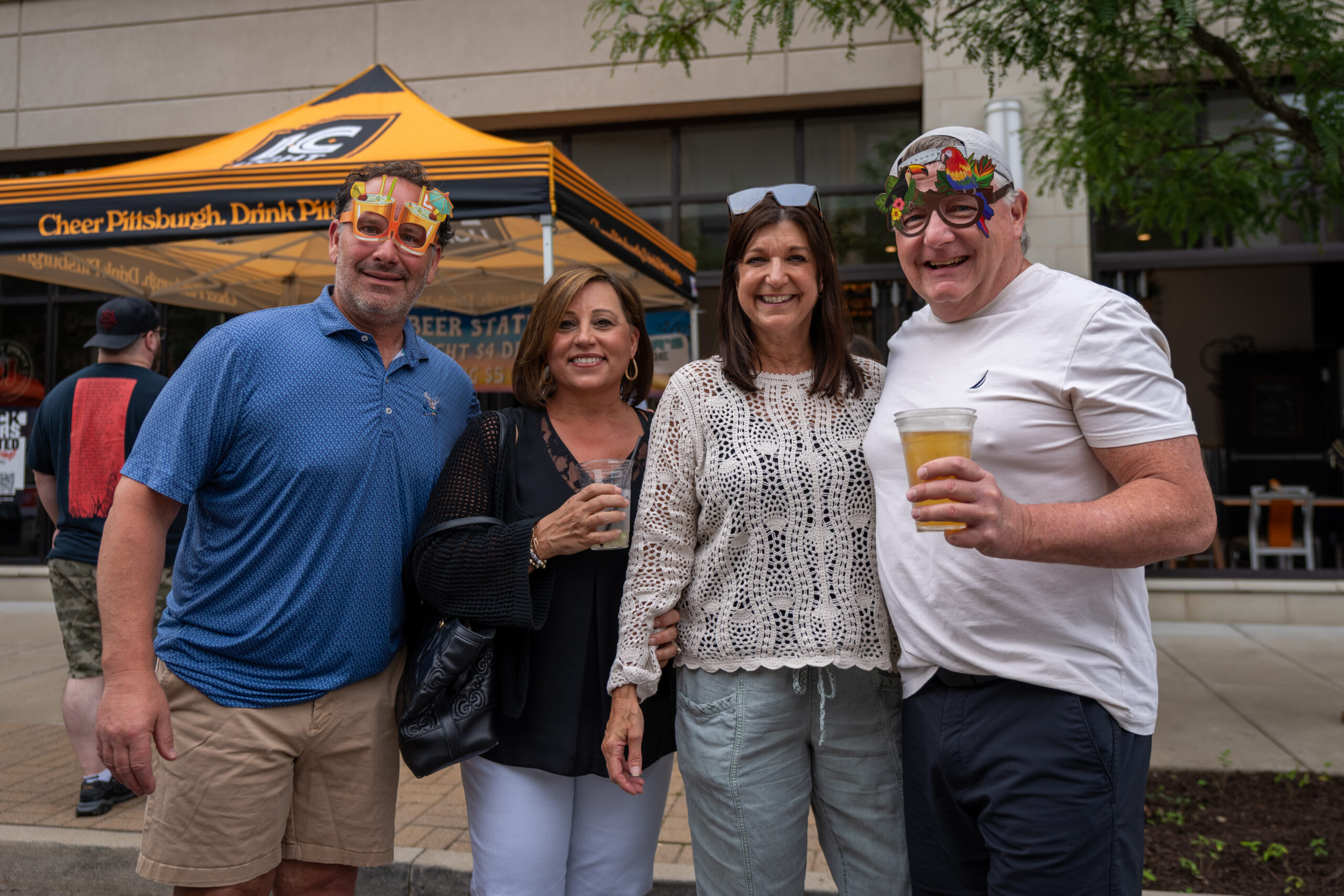 Four adults pose and smile outdoors at a street event. Two men wear colorful, festive glasses and hold drinks. A yellow tent with text and other people can be seen in the background at Canonsburg Live Music festival.