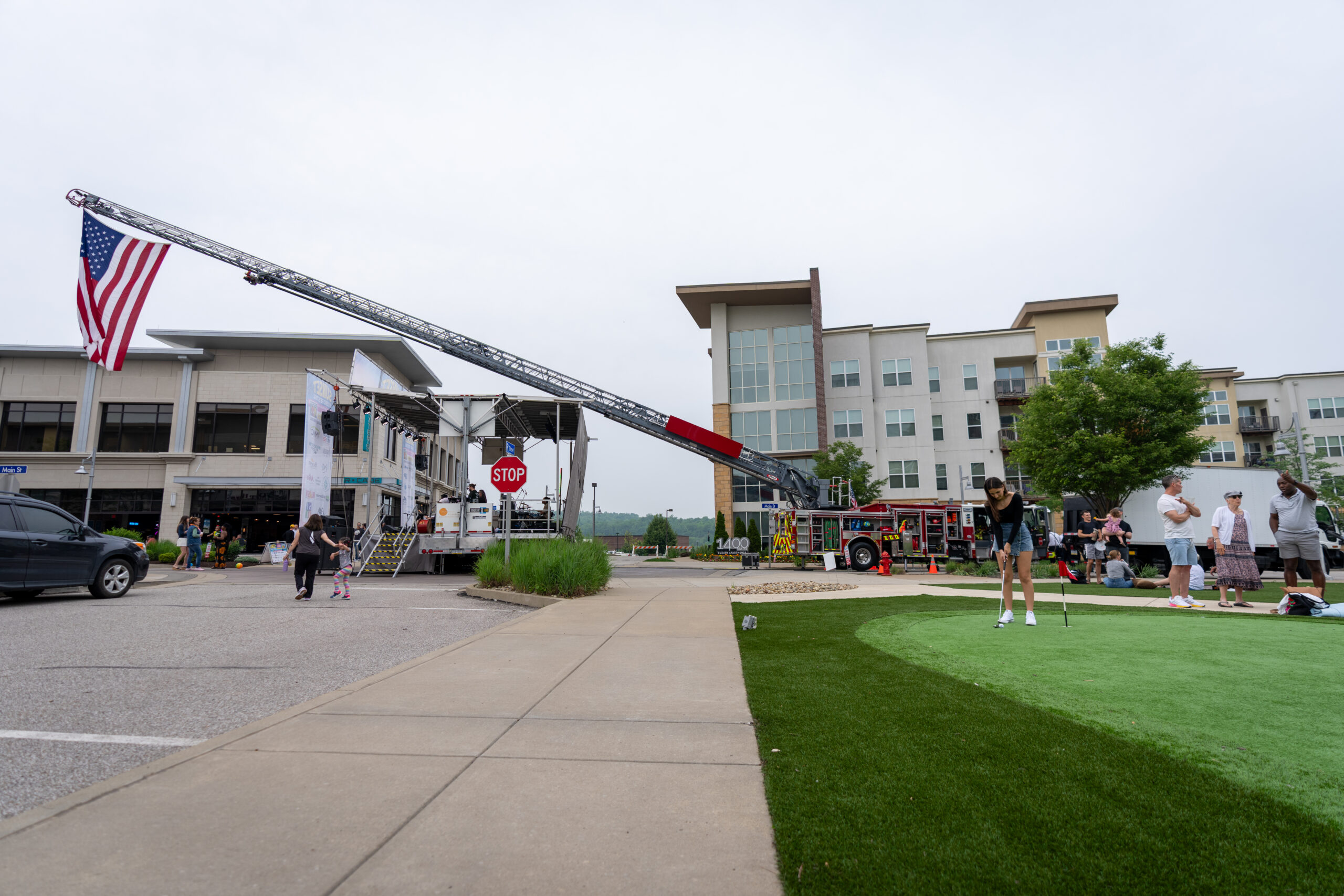 A fire truck extends its ladder displaying an American flag in front of an apartment building; people gather nearby, some playing mini-golf on a green lawn during the Canonsburg Live Music festival.