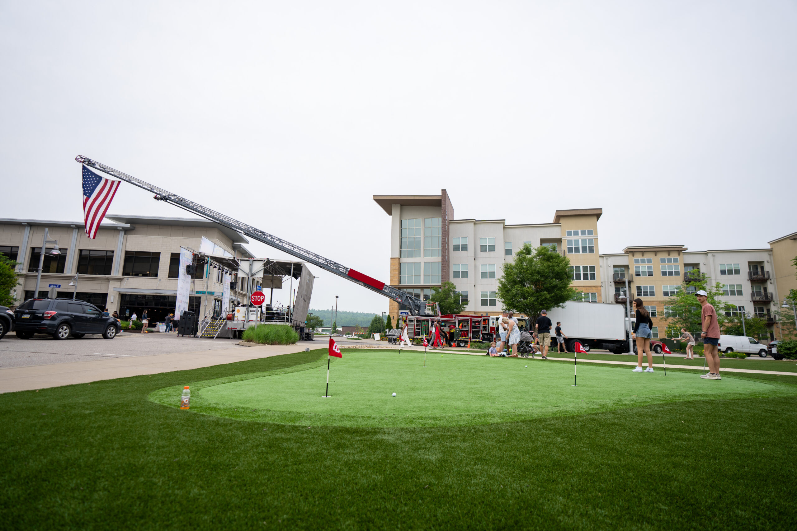 A small outdoor putting green with several people nearby, surrounded by modern buildings. During the Canonsburg Live Music event, a firetruck extends a ladder displaying a large American flag in the background beneath an overcast sky.