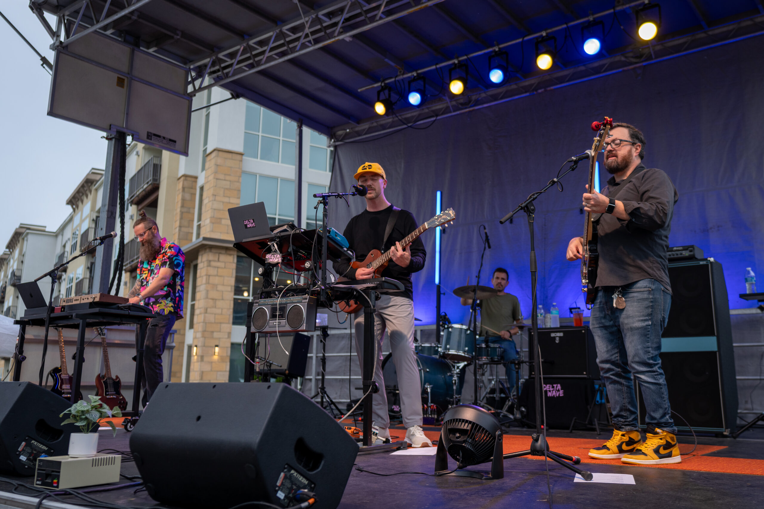 Four musicians perform on an outdoor stage at the Sunset Music Fest, playing keyboards, guitar, and drums. Blue and yellow lights illuminate the scene, with modern buildings visible in the background.