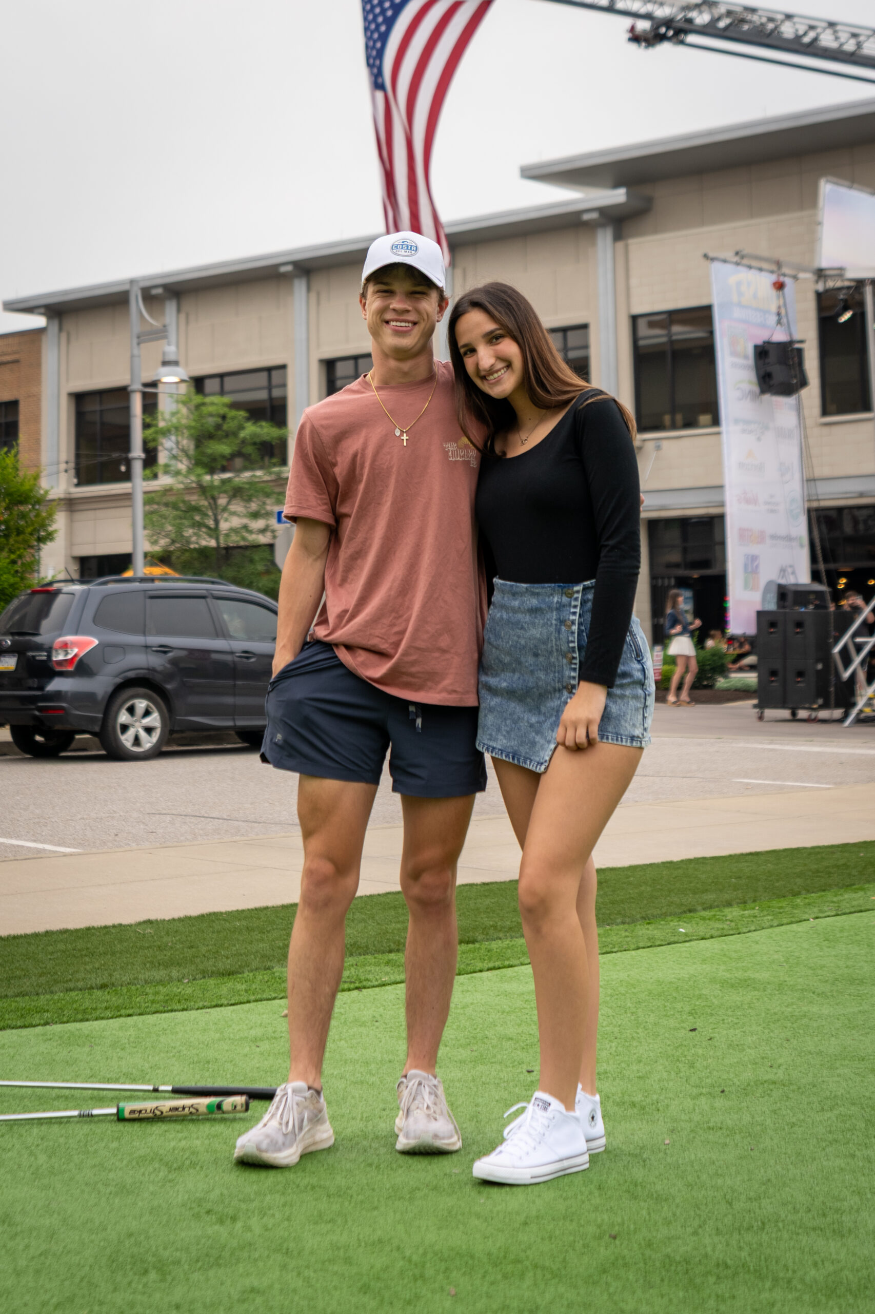 A young man in a pink shirt and shorts stands next to a young woman in a black top and denim skirt, both smiling on green artificial grass at the Sunset Music Fest with an American flag and buildings in the background.
