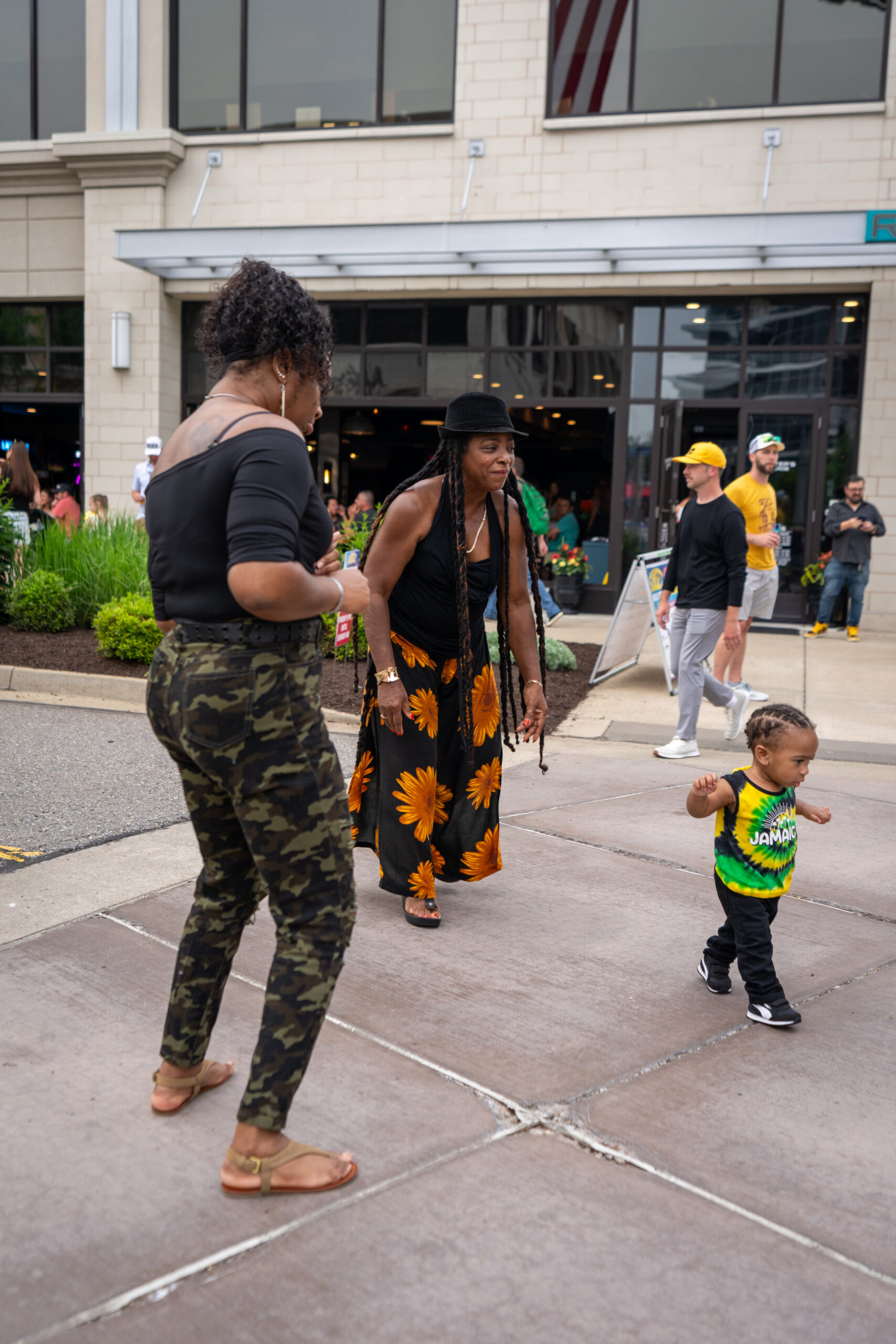 Two women smile and watch a toddler walk outdoors on a sidewalk, dressed for the Sunset Music Fest. One wears camouflage pants and a black top, the other a floral skirt and hat. People and a building are visible in the background.