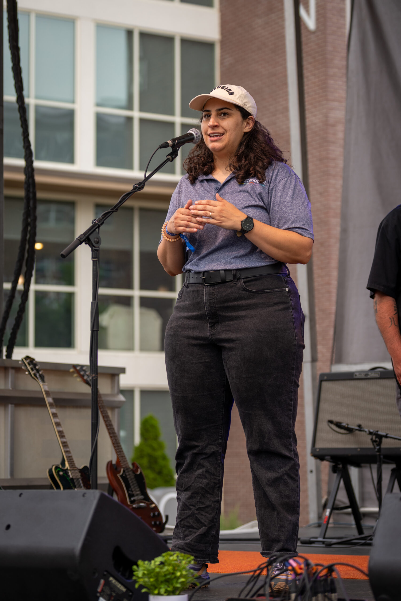 A person wearing a light-colored cap, blue polo shirt, and black pants speaks into a microphone on an outdoor stage at Canonsburg Live Music. Guitars, amplifiers, and buildings can be seen in the background.