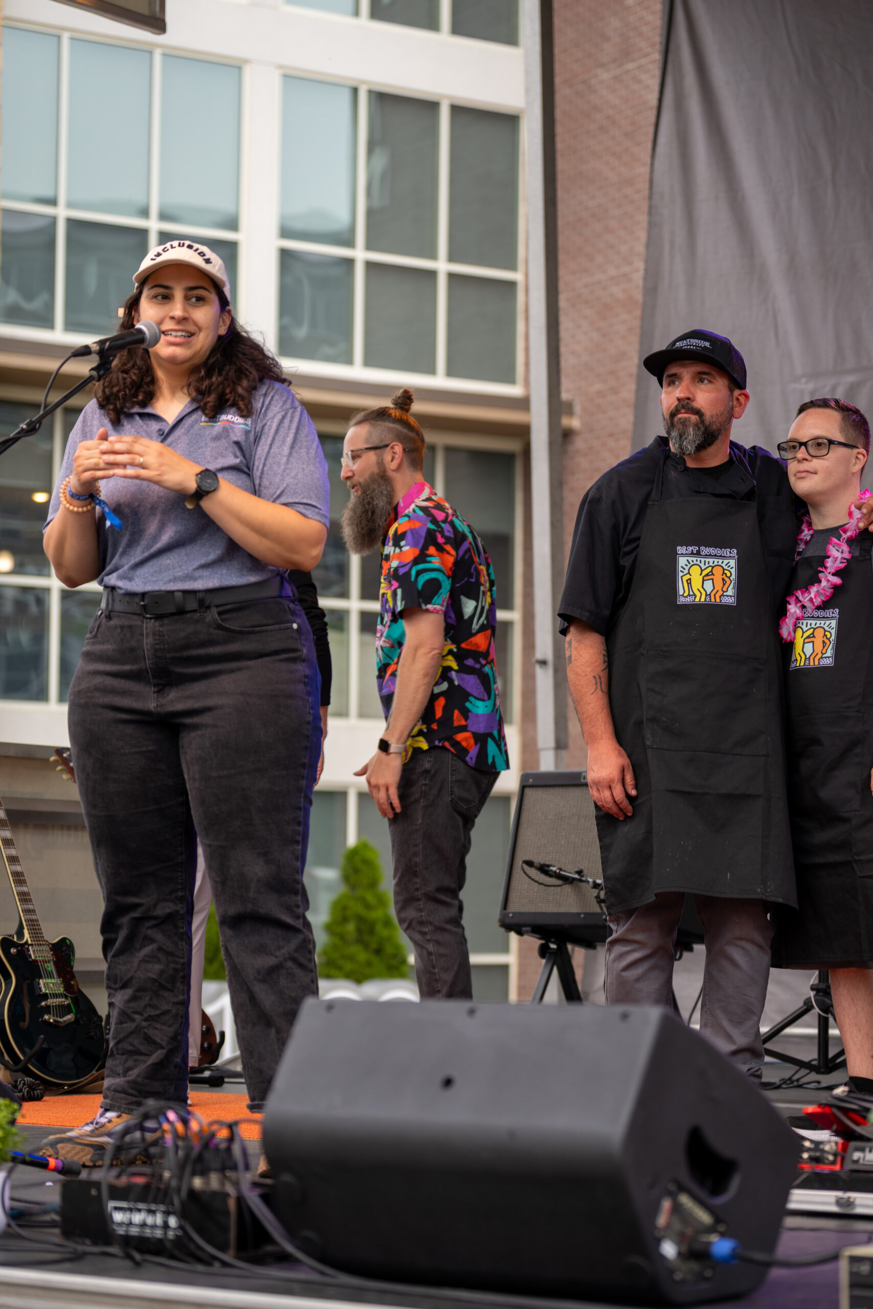 A woman speaks at a microphone on an outdoor stage during the Sunset Music Fest while three people, two in aprons, stand nearby. Musical equipment and a building with large windows are visible in the background.