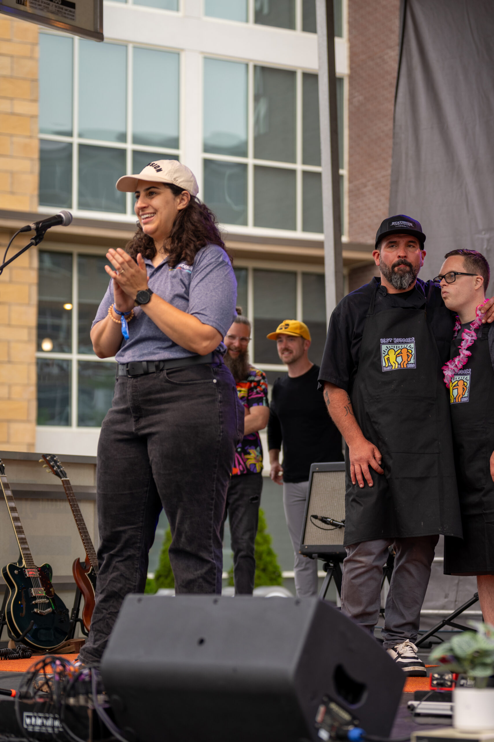 A woman in a white cap claps and smiles on an outdoor stage with a microphone during the Canonsburg Live Music festival. Behind her, four men stand—two in black aprons, one in a colorful shirt and yellow hat—with musical instruments nearby.
