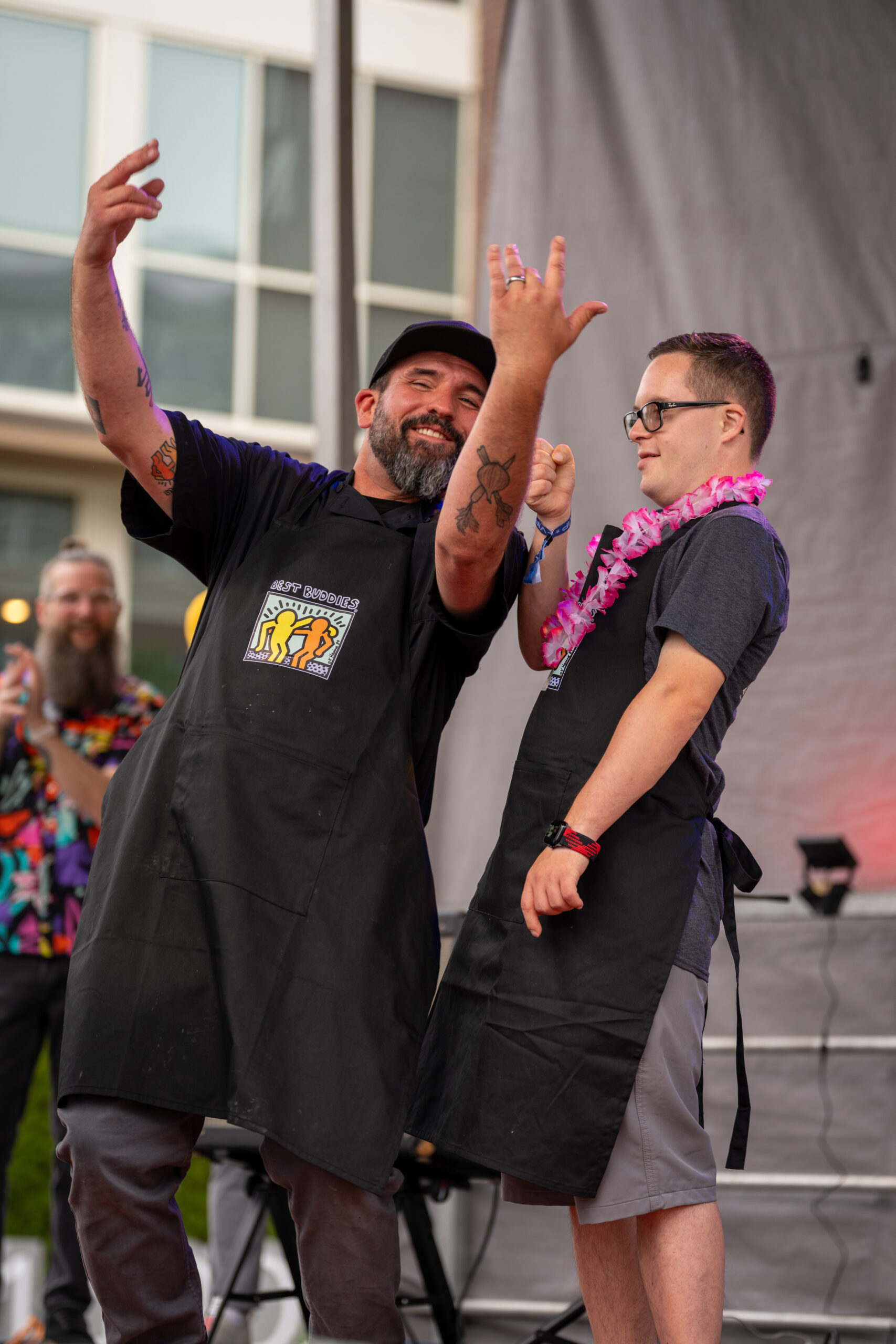 Two men wearing black aprons stand on an outdoor stage at Sunset Music Fest, smiling and gesturing playfully. One sports a cap and flexes his arms, while the other wears glasses and a pink lei. People and a building linger in the background.