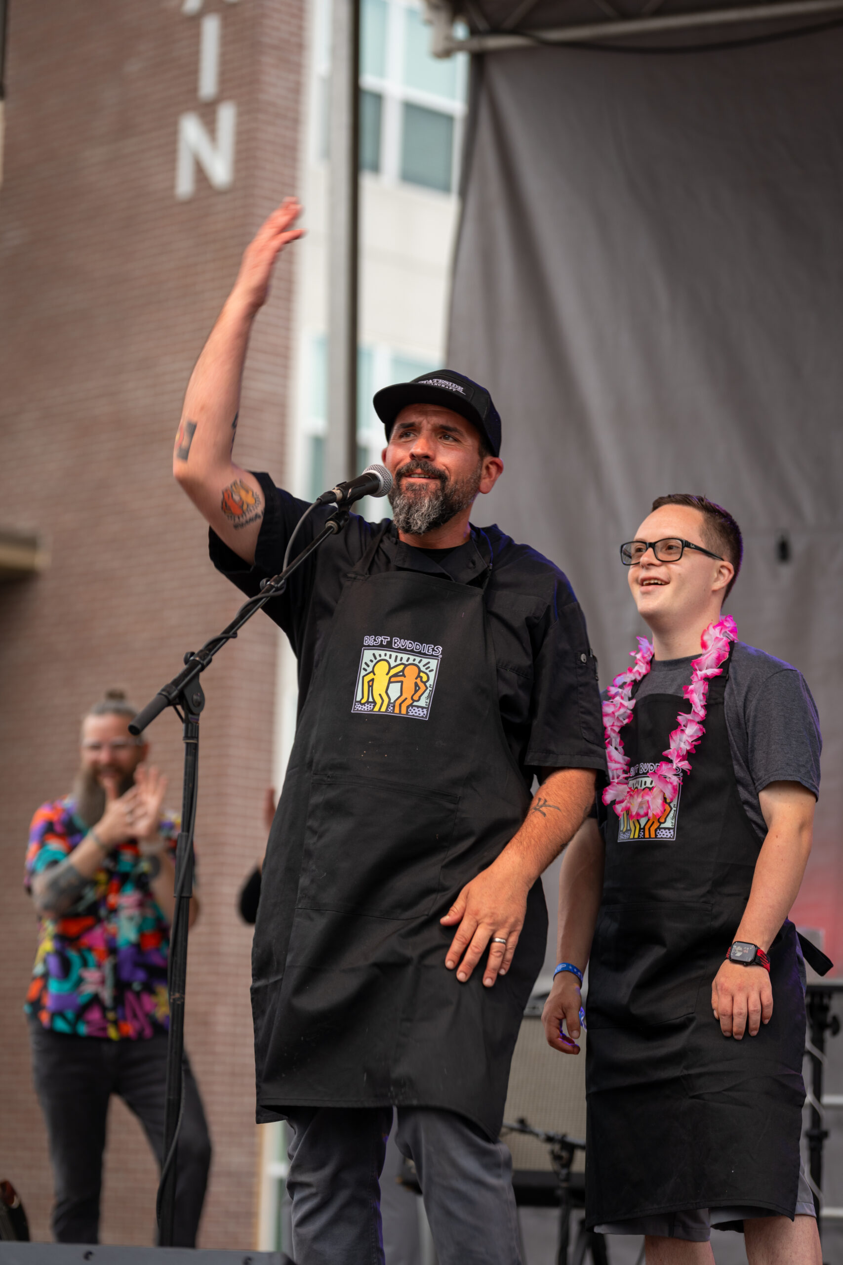 Two men in black aprons, one with a cap and raised arm, stand on an outdoor stage with microphones at Sunset Music Fest. The man beside him wears glasses and a pink lei. People and a brick building can be seen in the background.