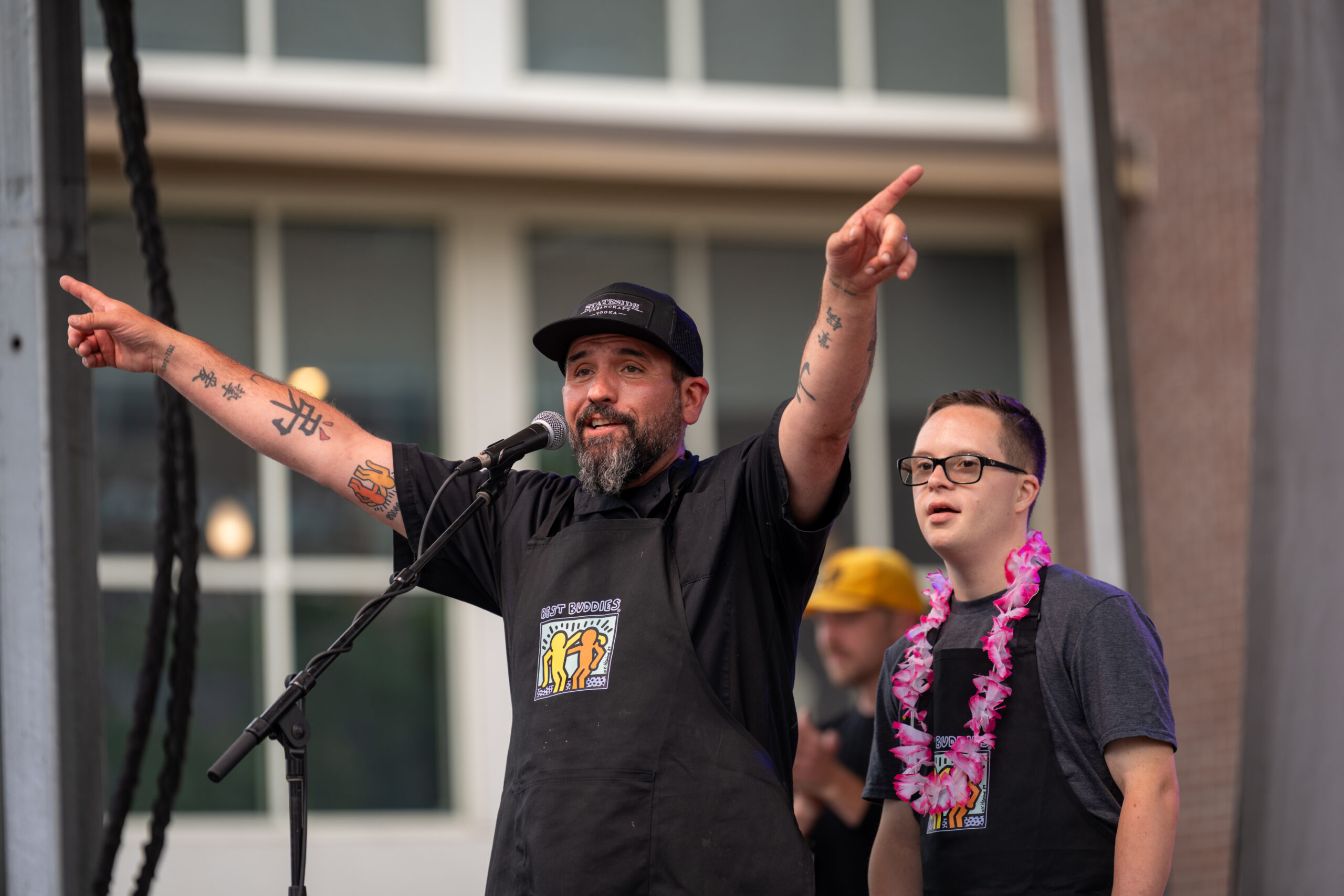 A man with tattoos speaks excitedly into a microphone with both arms raised, while another man in glasses and a pink lei stands beside him. Both wear black aprons on an outdoor stage at the Sunset Music Fest.