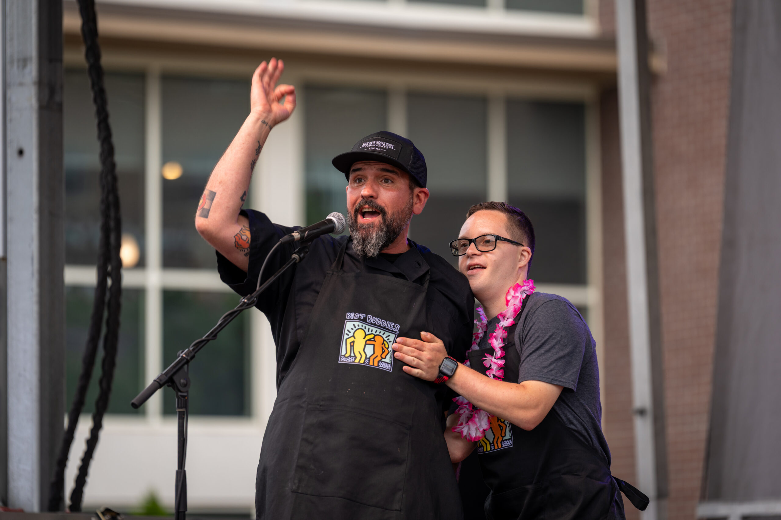 Two men wearing black aprons stand on stage at the Sunset Music Fest. One speaks enthusiastically into a microphone, arm raised, while the other stands beside him smiling, hand on his shoulder and wearing a pink lei.