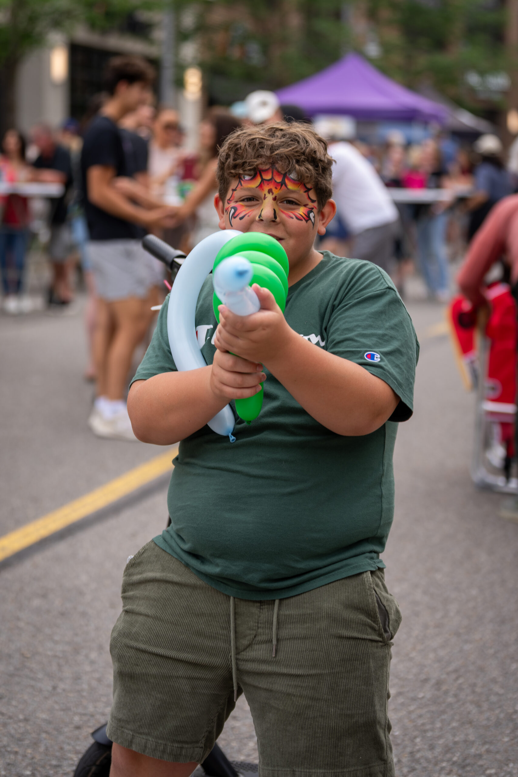 A child with face paint and glasses holds a green and white balloon animal while standing on a street at Sunset Music Fest; people and tents are visible in the blurred background at this lively outdoor event.