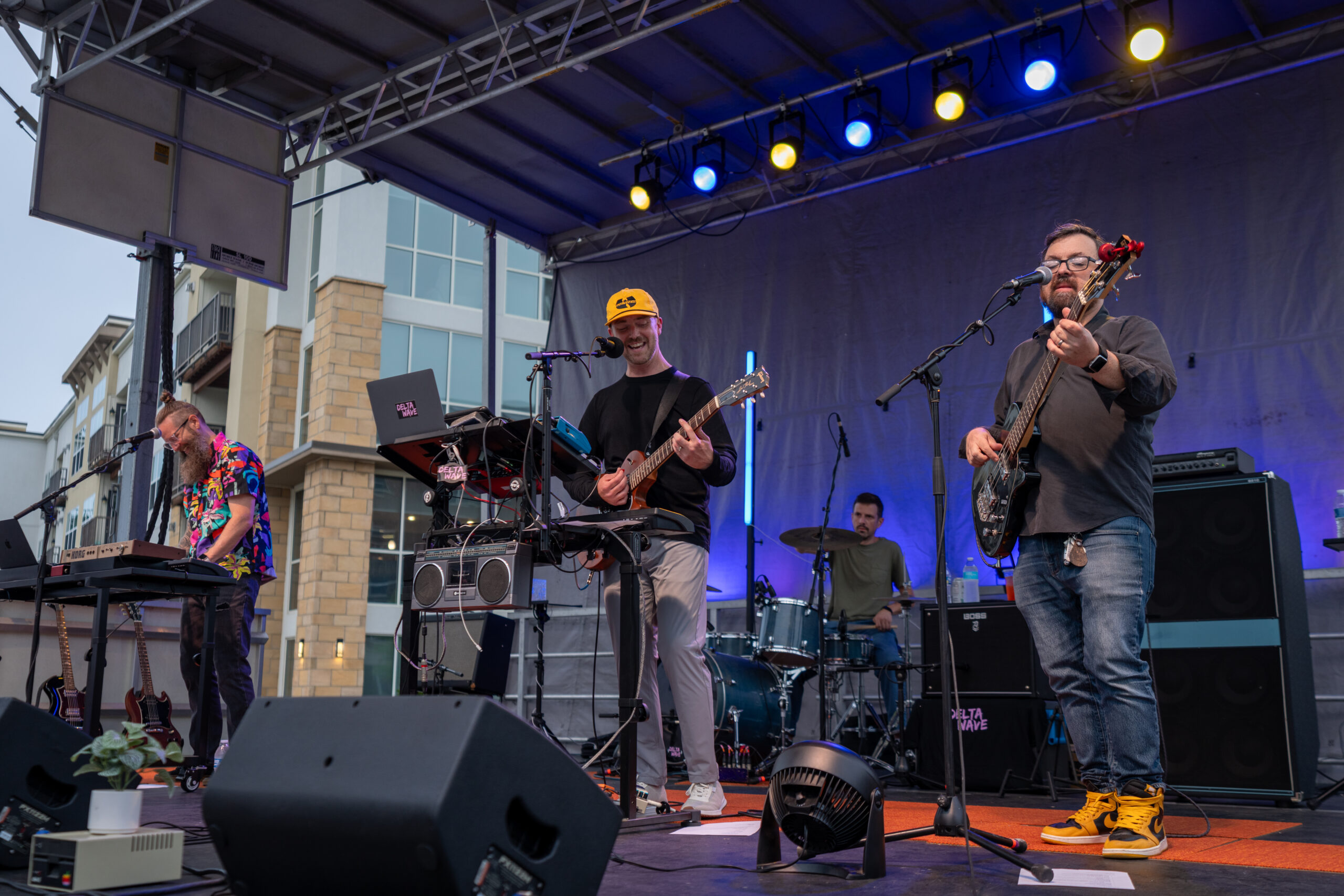 A four-member band performs on an outdoor stage at dusk during the Sunset Music Fest, playing guitars, keyboard, and drums under blue and yellow lights. The musicians are focused on their instruments, with buildings visible in the background.