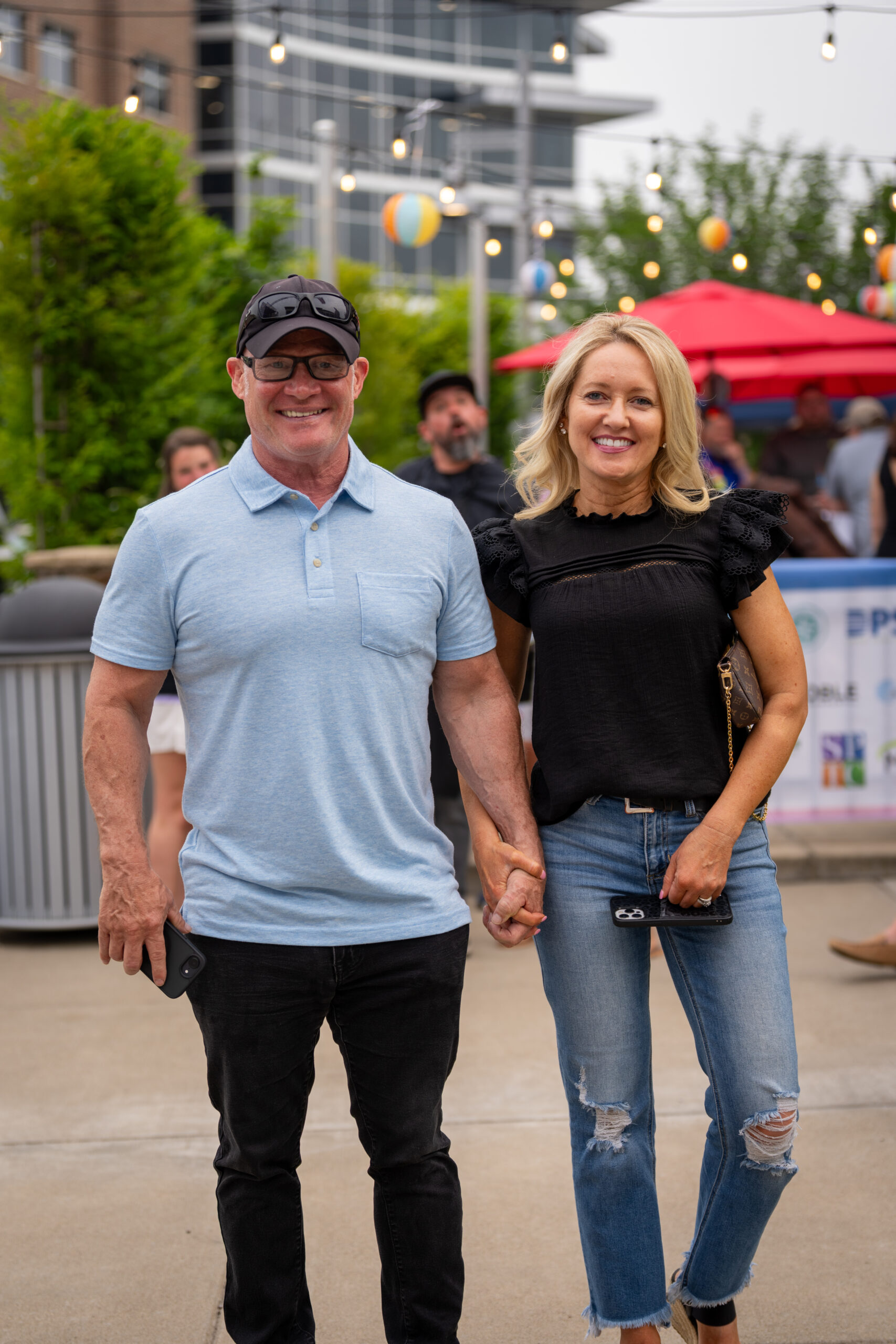 A smiling man in a light blue polo and black cap holds hands with a woman in a black blouse and ripped jeans as they walk outdoors at Sunset Music Fest, with string lights and fellow music festival guests in the background.