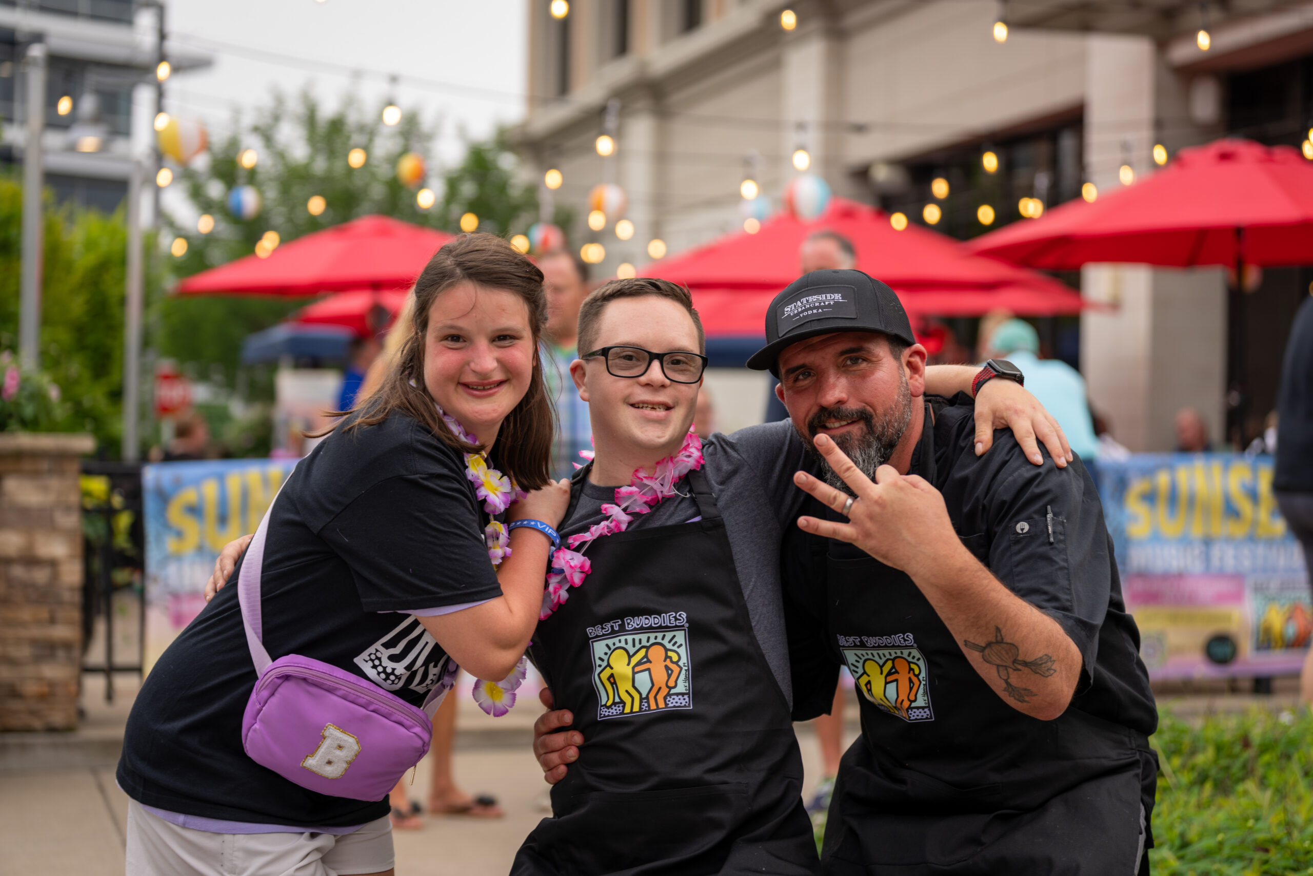 Three people pose and smile outdoors at the Sunset Music Fest. Two young individuals wear matching shirts and leis, while an adult in a chef’s hat gestures with three fingers. Red umbrellas and string lights set a festive background.