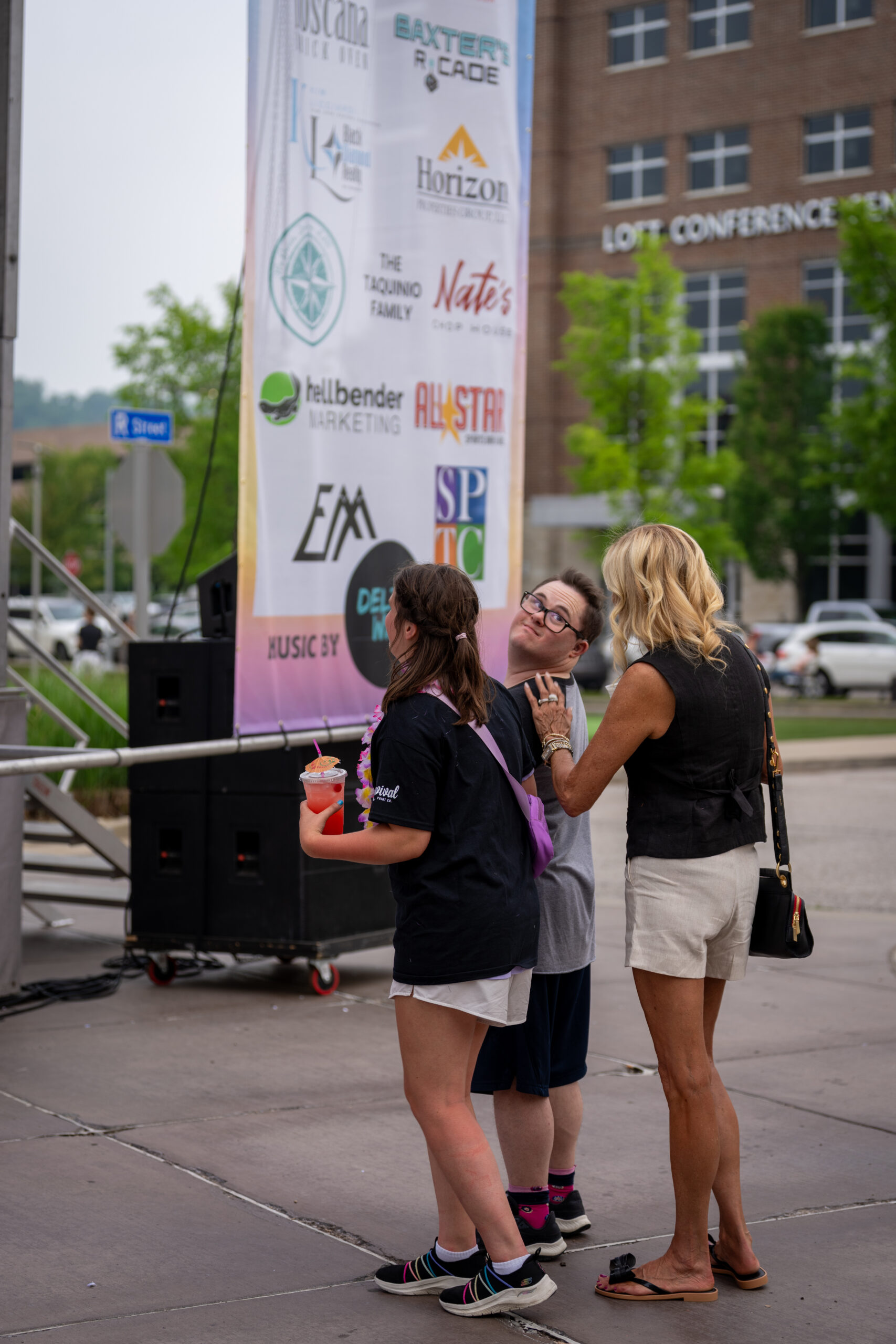 Three people stand together outdoors near a large event banner at Sunset Music Fest. Two appear to be younger and one adult; all are smiling and looking up, with one holding a drink. Cars and buildings are visible in the background.