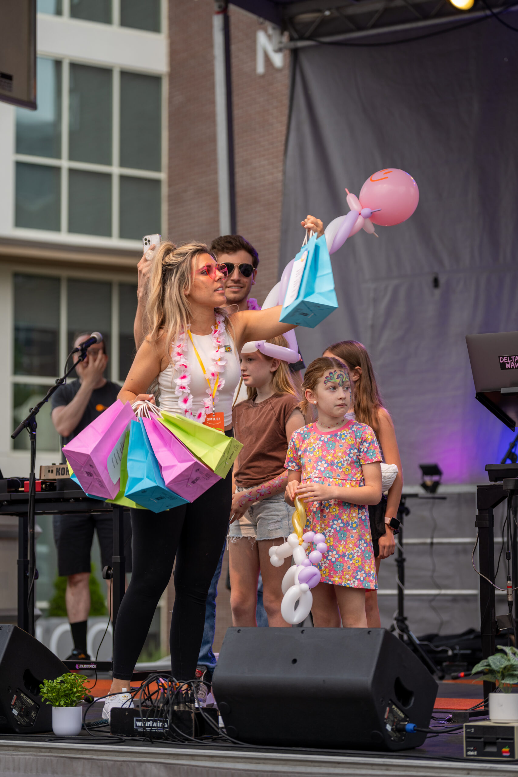 A woman on stage at the Sunset Music Fest holds colorful gift bags and a pink balloon animal, while children with painted faces and balloon creations stand nearby. Musical equipment and a keyboard are visible in the background.