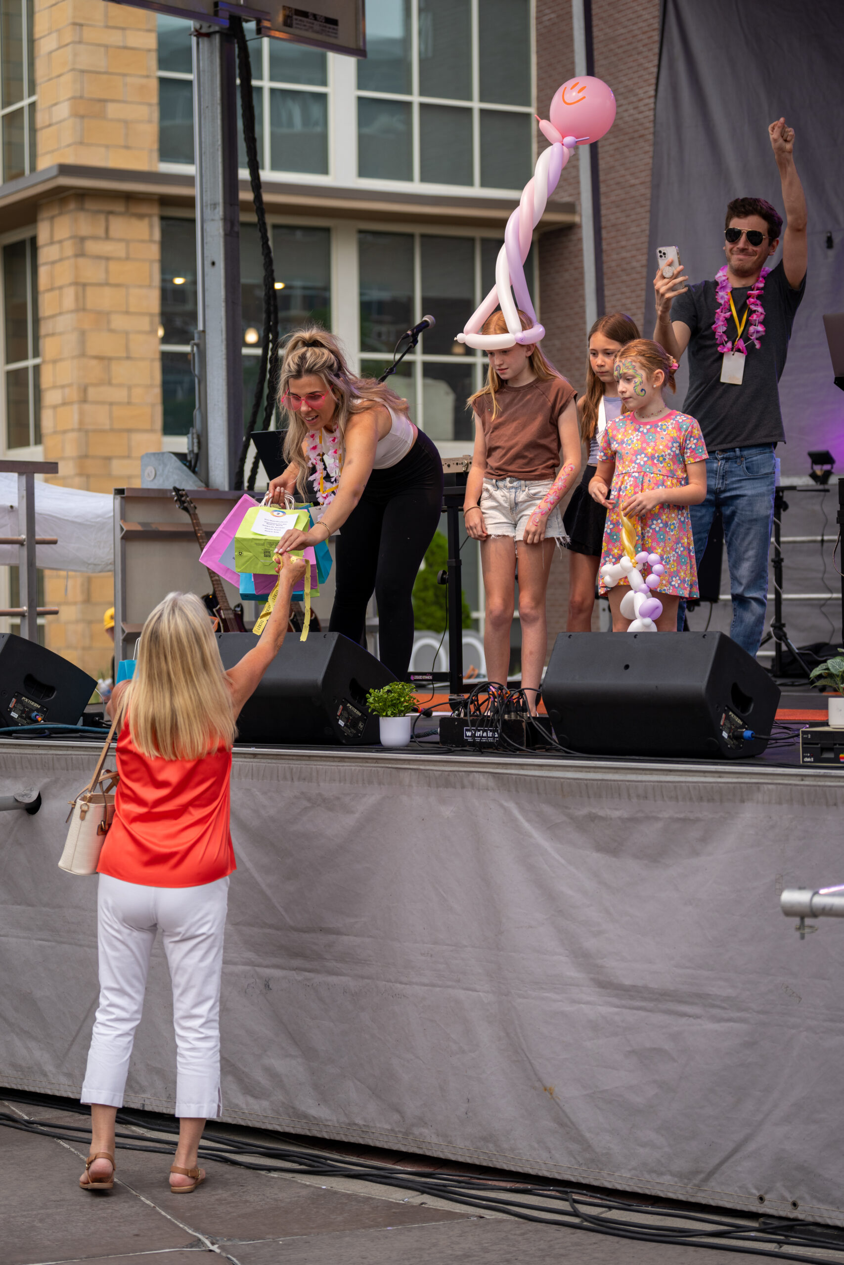 A woman on stage hands a colorful gift bag to an audience member as three children, one in a large balloon hat, stand nearby. A man in sunglasses applauds while the crowd enjoys the lively exchange at the Sunset Music Fest.